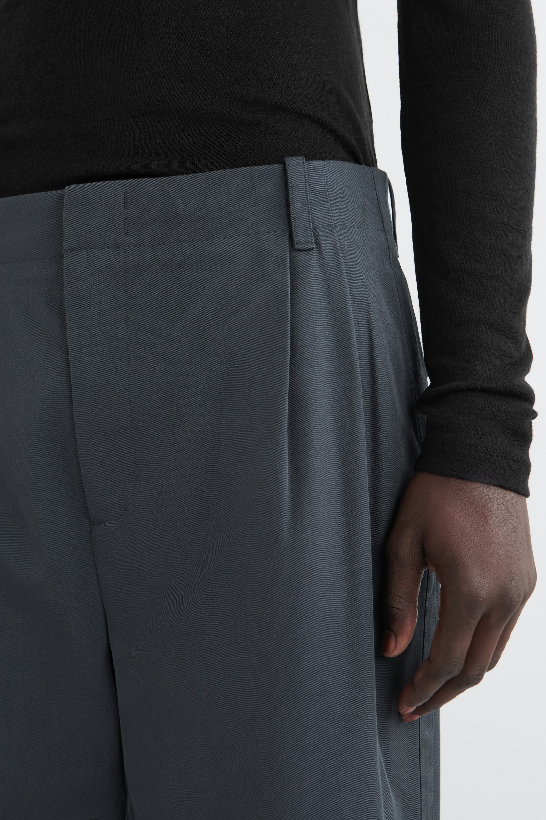 View larger image: Dark gray trousers are being presented from a front-right angle, featuring a flat waistband with a visible belt loop. A single pleat is falling from the waist, and a black long-sleeved top is being tucked into the trousers, with a hand resting on the side.
