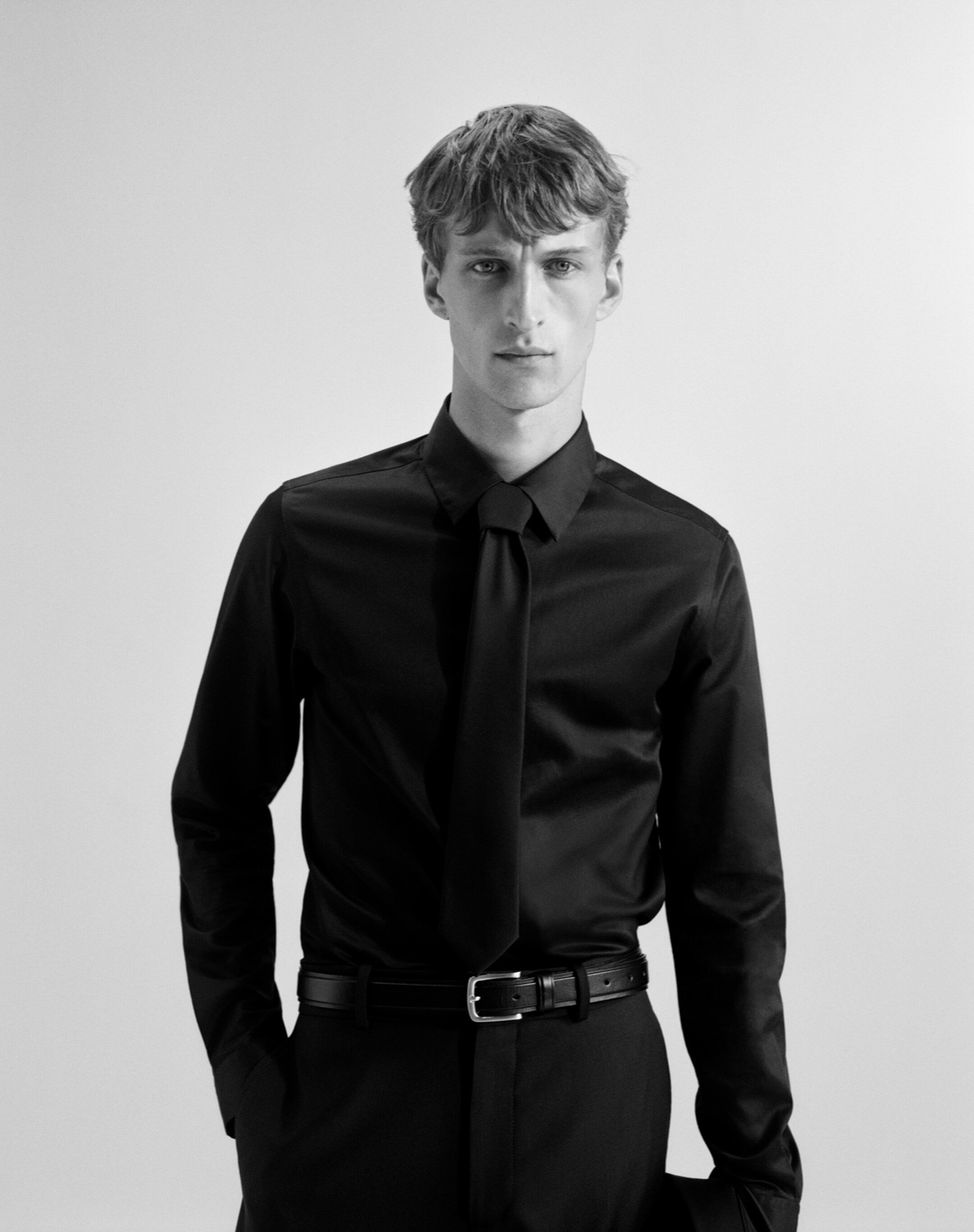 Größeres Bild anzeigen: Ein schwarzes langärmliges Hemd wird von einem Modell getragen, das nach vorne schaut. Das Slim-Fit-Hemd verfügt über einen spitzen Kragen, einen Knopfverschluss und lange Ärmel mit Manschetten und ist mit einer schwarzen Krawatte gestylt und in schwarze Hosen gesteckt.