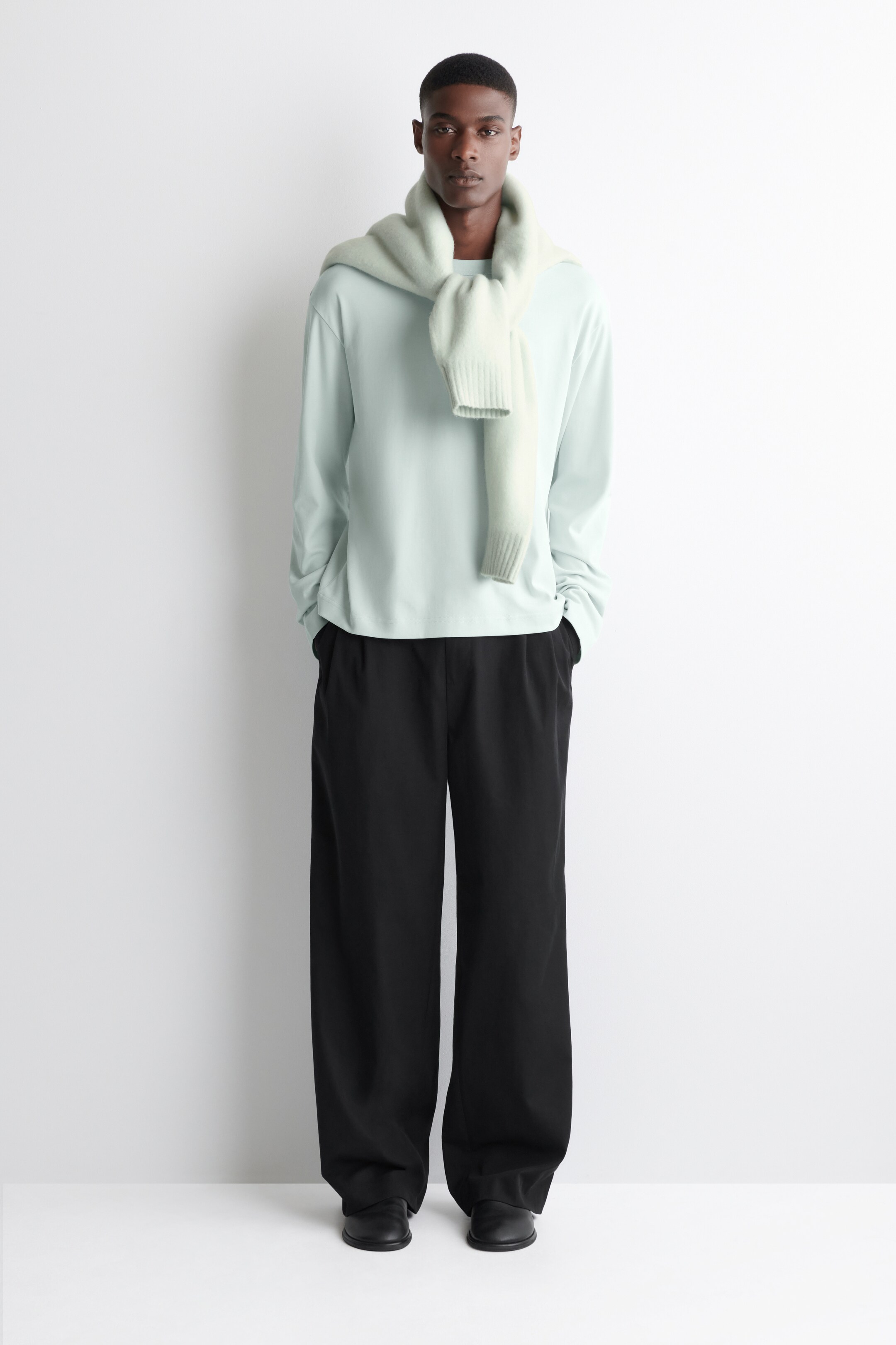 View larger image: A relaxed-fit, light mint long-sleeve crewneck top is being worn with black wide-leg trousers and black shoes, presented from a front-facing angle. A light green chunky knit sweater is being draped over the shoulders, resting on the top, with ribbed cuffs visible.