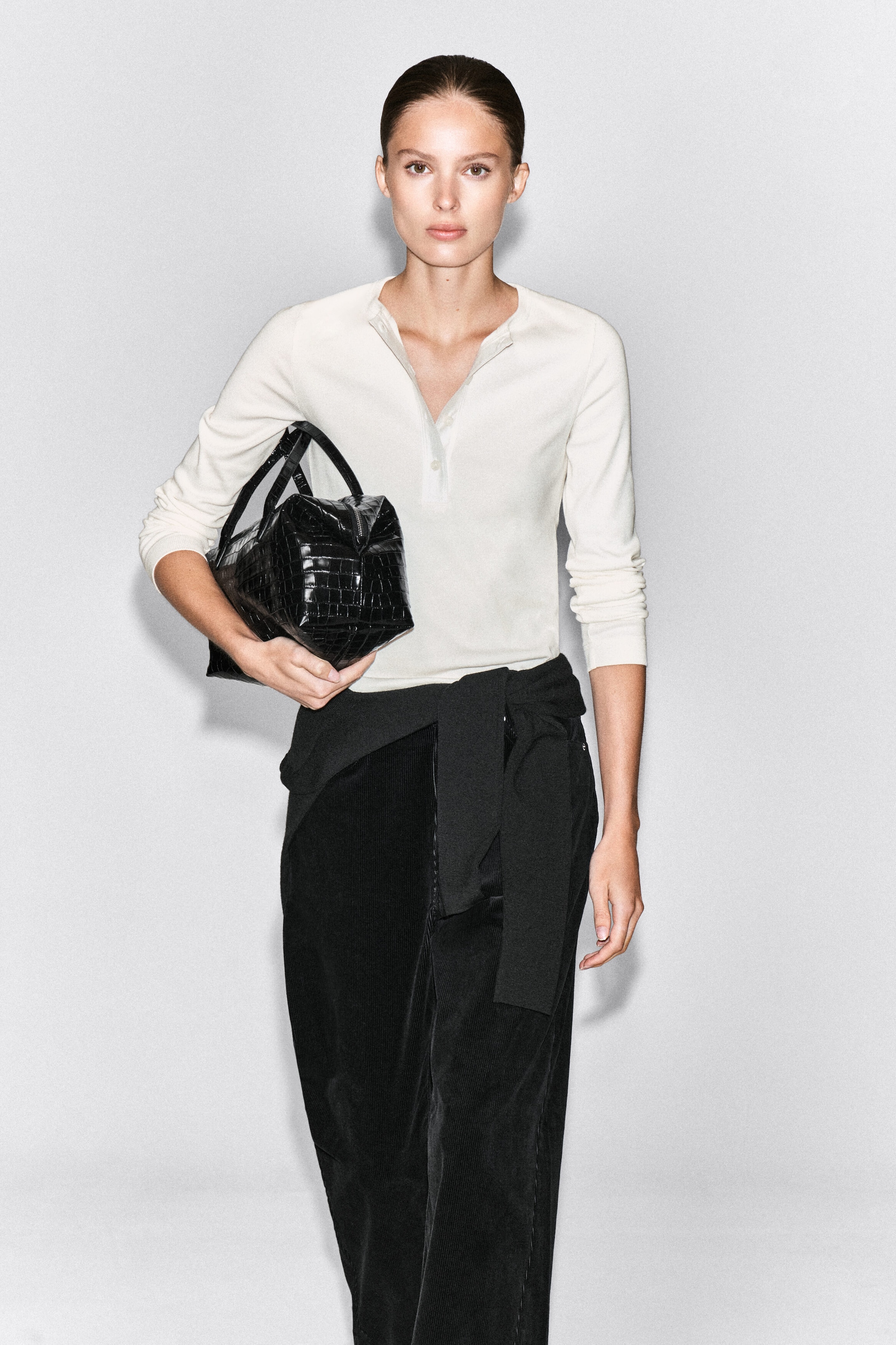 The model is wearing an off-white ribbed cotton Henley top with a partially unbuttoned collar and long sleeves. It is styled with black trousers, a black sweater tied at the waist, and a black textured handbag carried by the model. The model is wearing an off-white ribbed cotton Henley top with a partially unbuttoned collar and long sleeves. It is styled with black trousers, a black sweater tied at the waist, and a black textured handbag carried by the model.