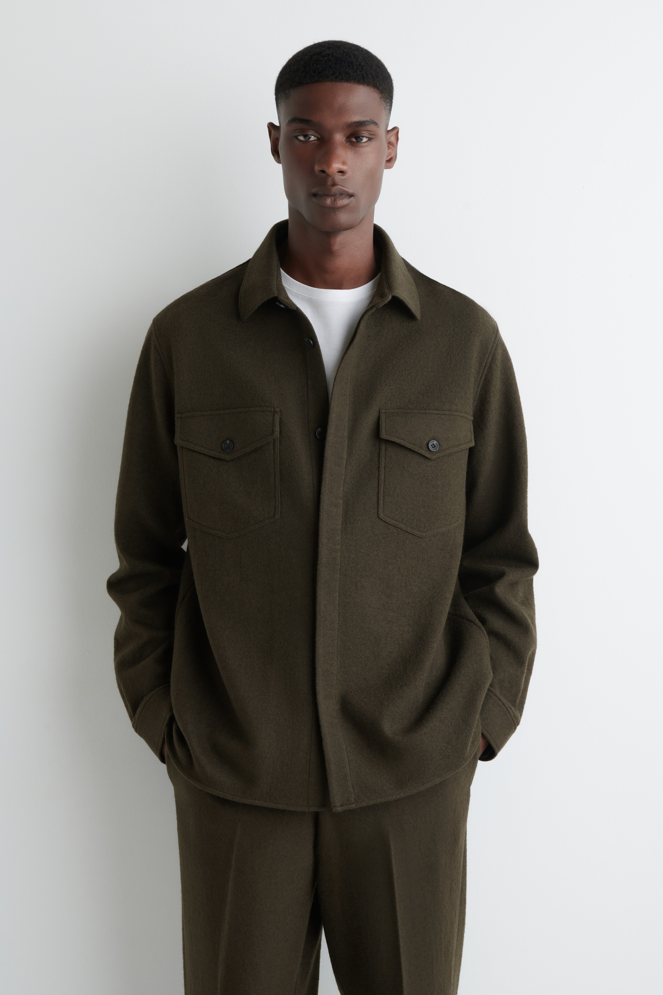 Agrandir l'image: Une surchemise vert olive en tissu brossé est présentée de face, avec un col classique en pointe et une fermeture à boutons sur le devant. Elle affiche deux grandes poches poitrine à rabats boutonnés et est superposée à un t-shirt blanc à col rond, assortie à un pantalon vert olive.