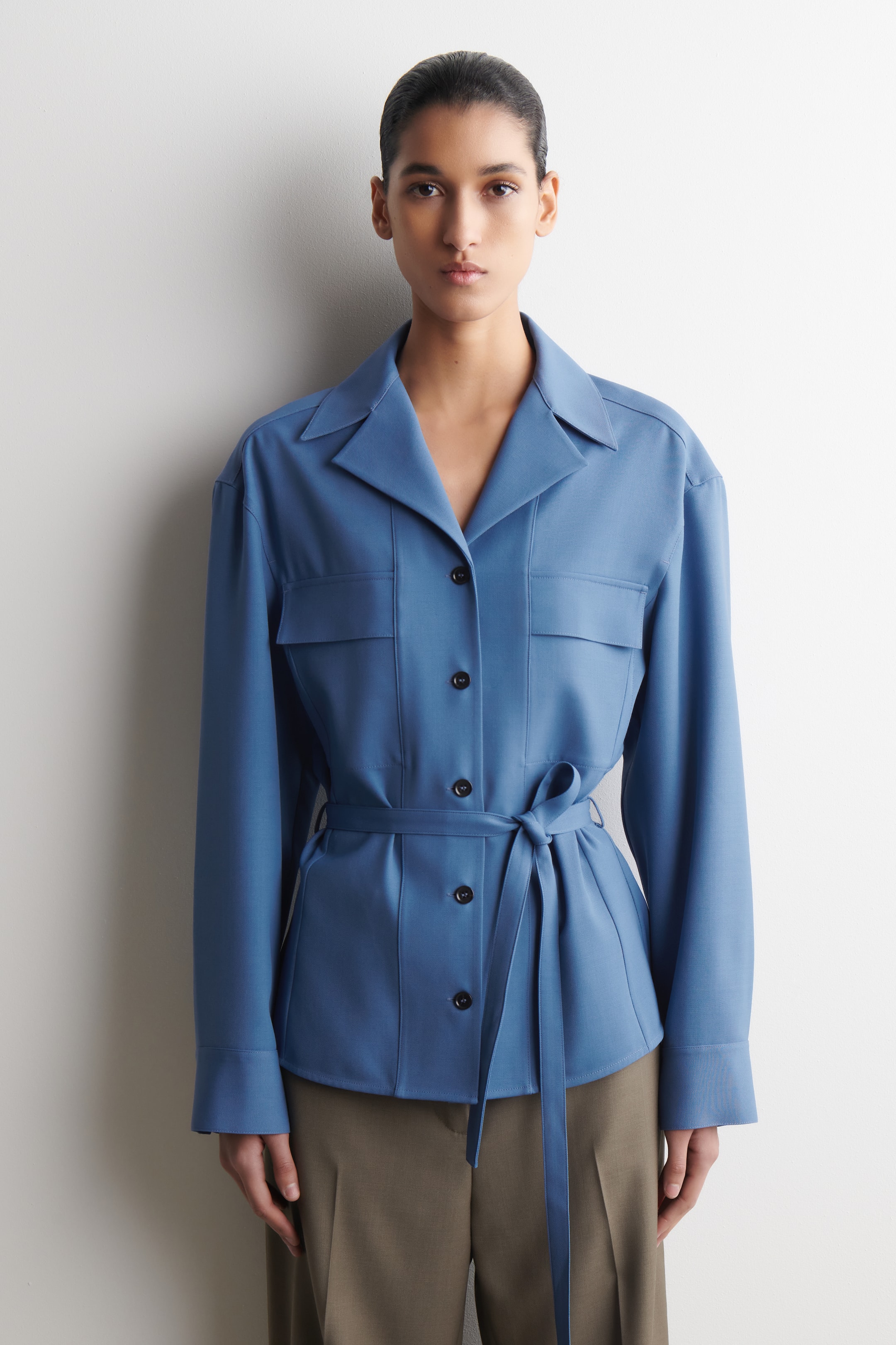 Προβολή μεγαλύτερης εικόνας: Ένα μπλε πουκάμισο-σακάκι διαθέτει γιακά με άνοιγμα, κλείσιμο μπροστά με μαύρα κουμπιά και δύο τσέπες στο στήθος με καπάκι. Σφίγγει στη μέση με μια ζώνη από το ίδιο ύφασμα δεμένη σε κόμπο και έχει μακριά μανίκια.