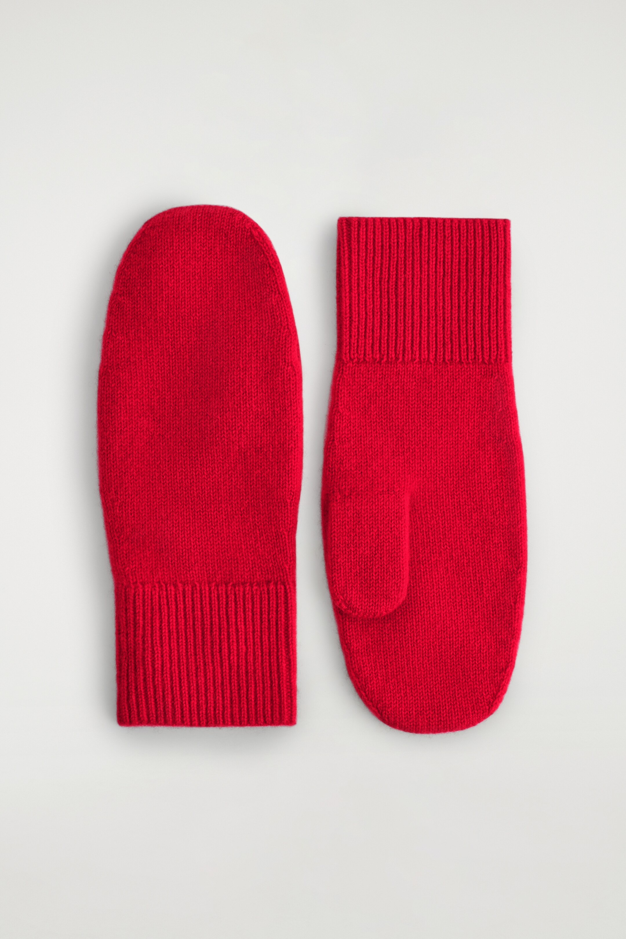View larger image: A pair of red knitted mittens is lying flat, featuring smooth main bodies and chunky ribbed cuffs, with one mitten displaying its individual thumb section.
