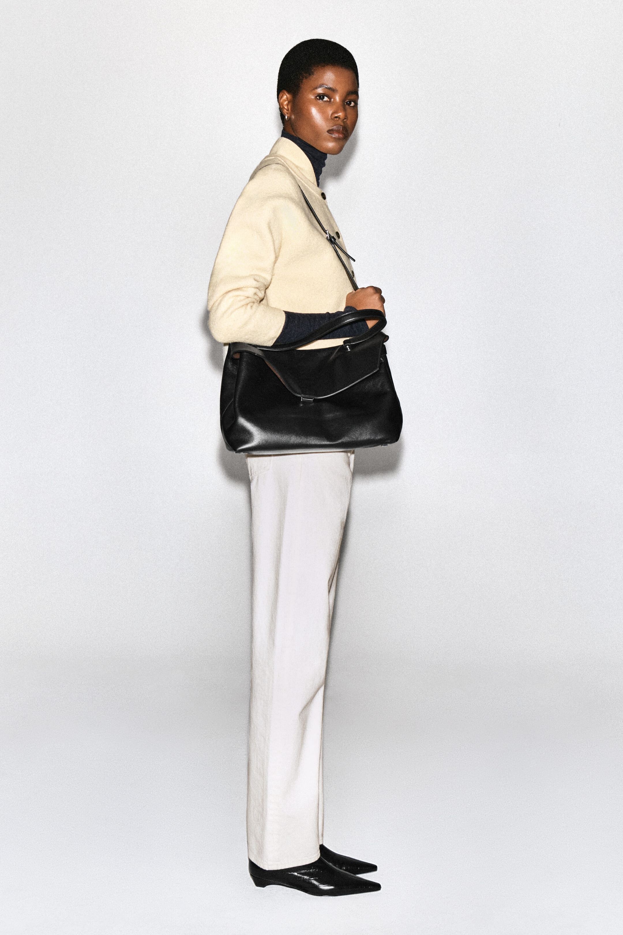 Un cardigan en laine bouillie couleur crème, à la silhouette arrondie et doté d’un col de style bomber, est porté par le mannequin par-dessus un col roulé foncé. Le mannequin pose de profil, assortissant le cardigan à un pantalon blanc et portant un sac à bandoulière en cuir noir. Un cardigan en laine bouillie couleur crème, à la silhouette arrondie et doté d’un col de style bomber, est porté par le mannequin par-dessus un col roulé foncé. Le mannequin pose de profil, assortissant le cardigan à un pantalon blanc et portant un sac à bandoulière en cuir noir.