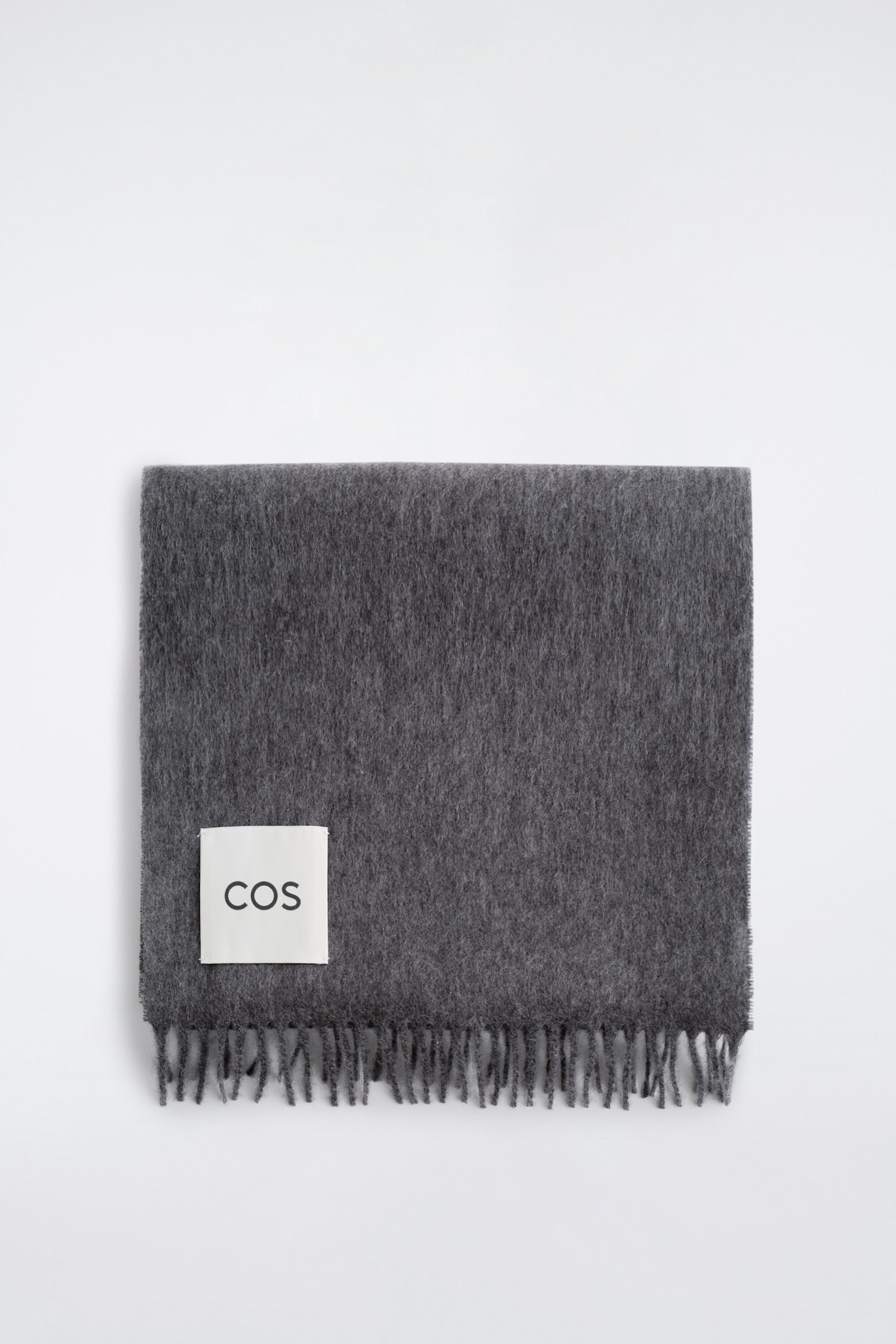 A dark grey fringed cashmere scarf is lying flat in a folded rectangular shape, showcasing its soft, brushed texture and fringed bottom edge. A square white label featuring the black "COS" brand name is stitched onto the lower left corner.