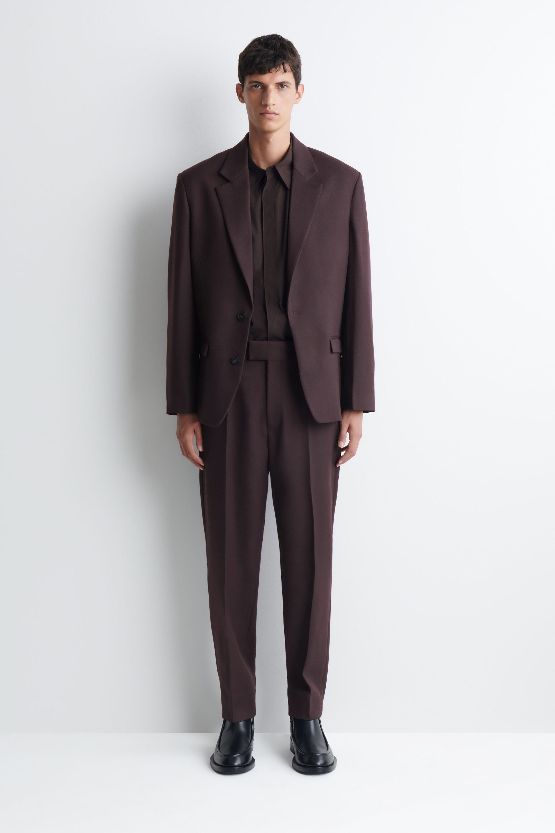 A dark brown suit, featuring an open single-breasted blazer with notched lapels and matching straight-leg trousers, is being worn by the model. A dark brown long-sleeve shirt is being layered underneath the blazer, and black boots are being worn with the trousers. A dark brown suit, featuring an open single-breasted blazer with notched lapels and matching straight-leg trousers, is being worn by the model. A dark brown long-sleeve shirt is being layered underneath the blazer, and black boots are being worn with the trousers.