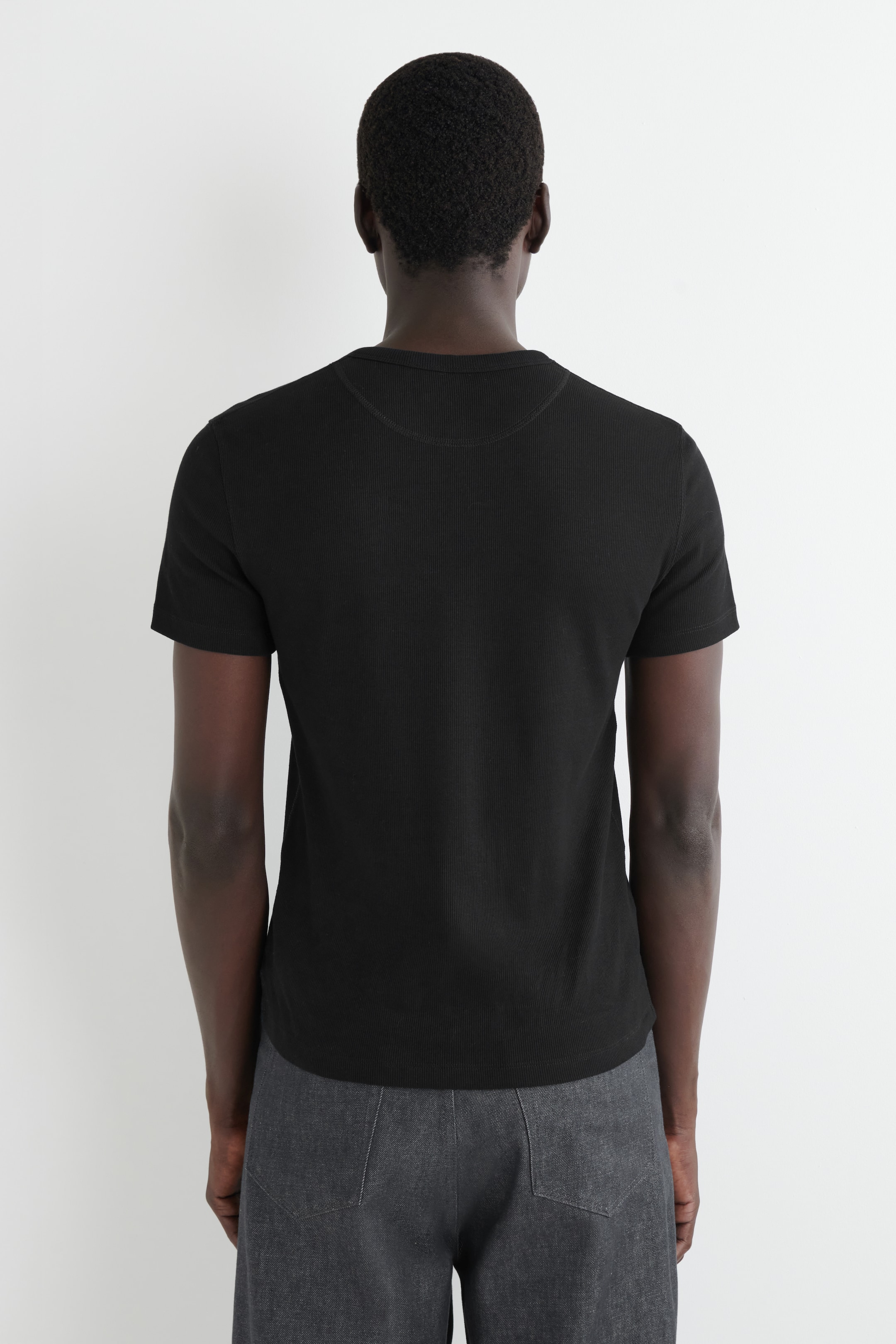 Agrandir l'image: Un t-shirt noir est présenté de dos, avec une texture côtelée et des manches courtes. Il est conçu avec un col rond, souligné par des coutures visibles le long de la bande du col.