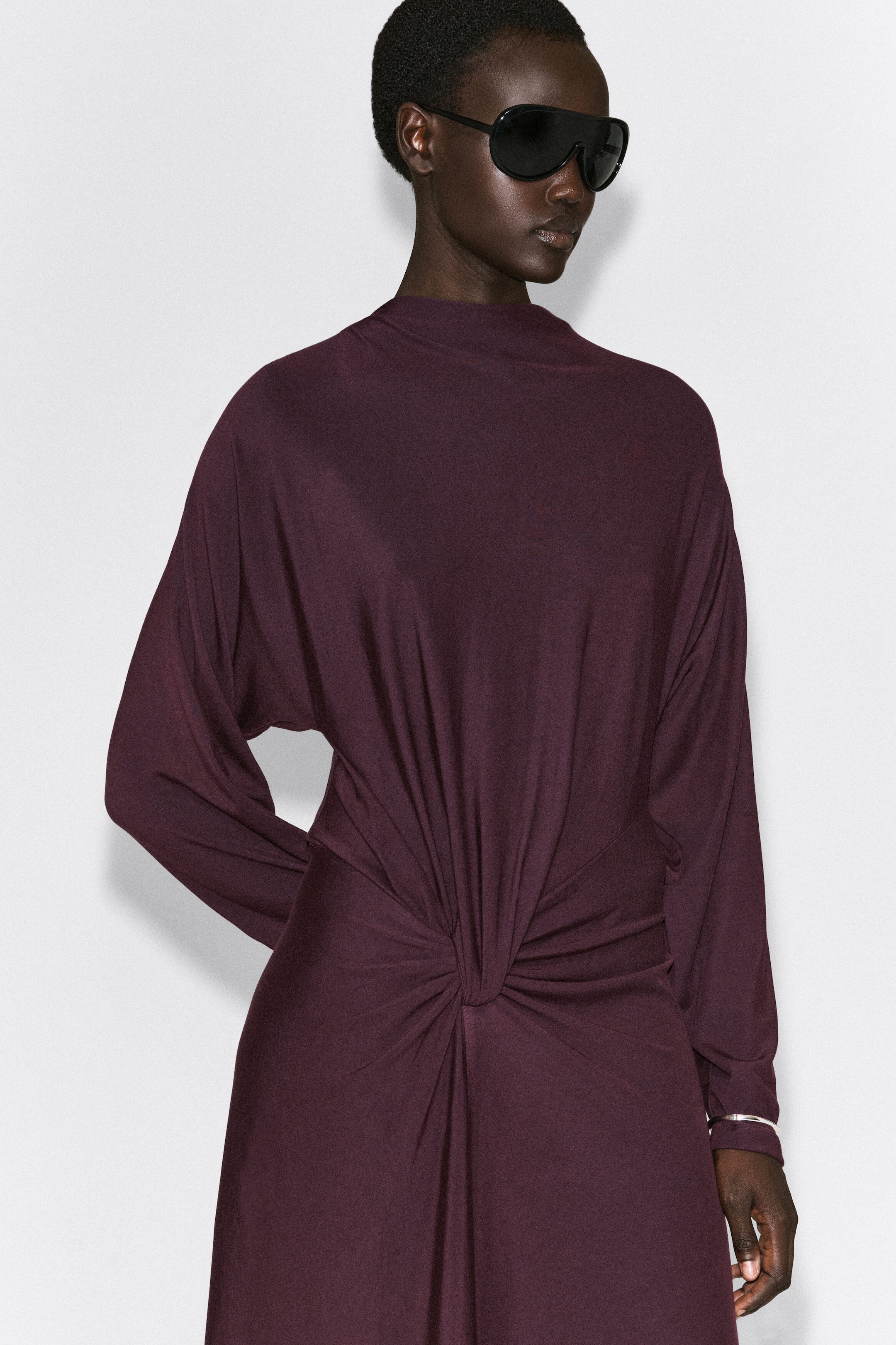 La robe midi drapée à torsade, couleur prune foncée, présentée de face, est dotée d’un col montant type mock neck et de manches longues amples. Un détail de nœud torsadé central à la taille rassemble le tissu, créant de doux plis drapés qui se déploient sur le corsage et la jupe. La robe midi drapée à torsade, couleur prune foncée, présentée de face, est dotée d’un col montant type mock neck et de manches longues amples. Un détail de nœud torsadé central à la taille rassemble le tissu, créant de doux plis drapés qui se déploient sur le corsage et la jupe.