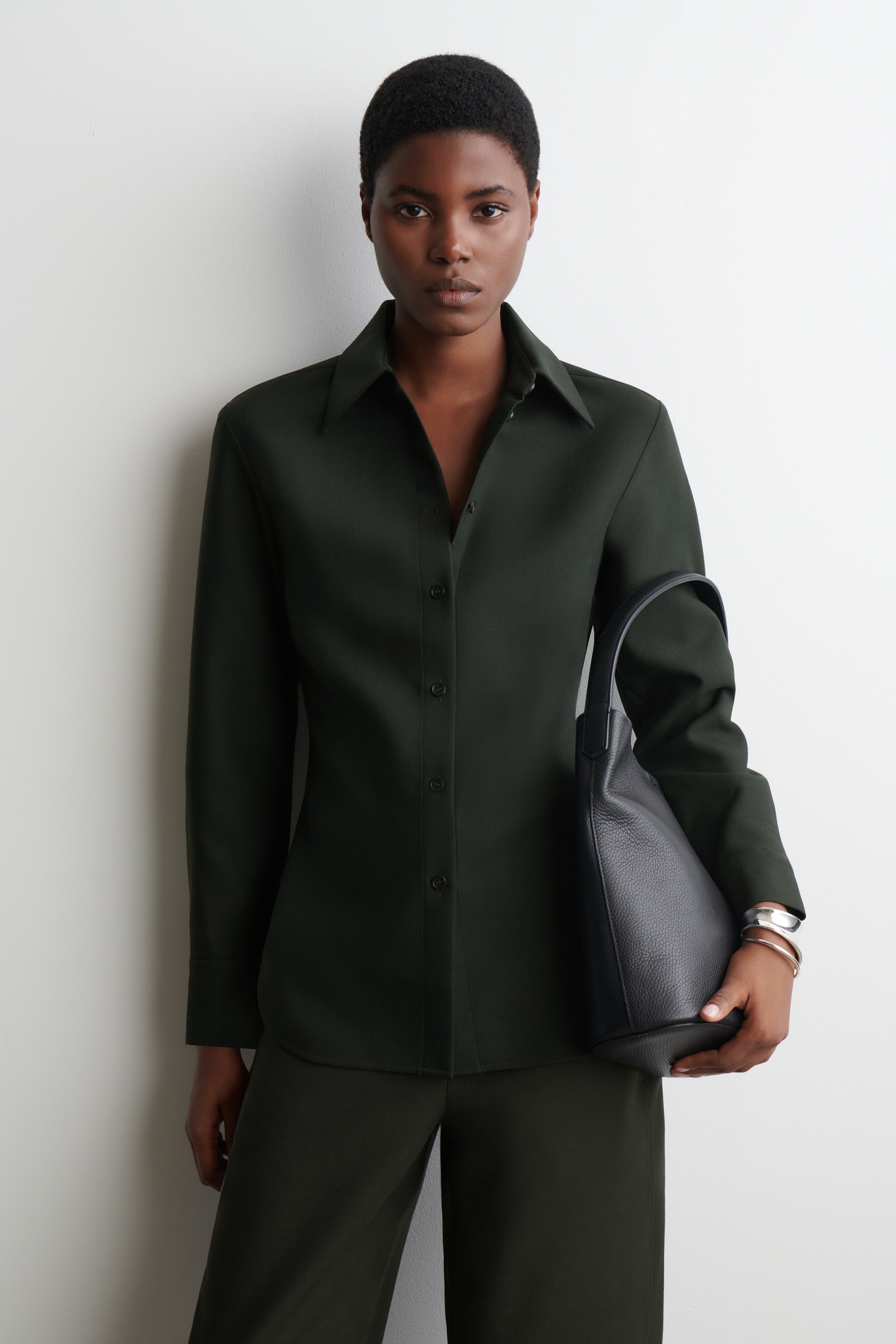 Agrandir l'image: Une chemise à manches longues vert foncé est présentée de face, avec un col pointu classique et une patte de boutonnage complète avec des boutons foncés assortis, mettant en valeur une silhouette ajustée. Un pantalon large vert foncé assorti est également porté. Un sac à main en cuir noir grainé avec une poignée supérieure est tenu devant.