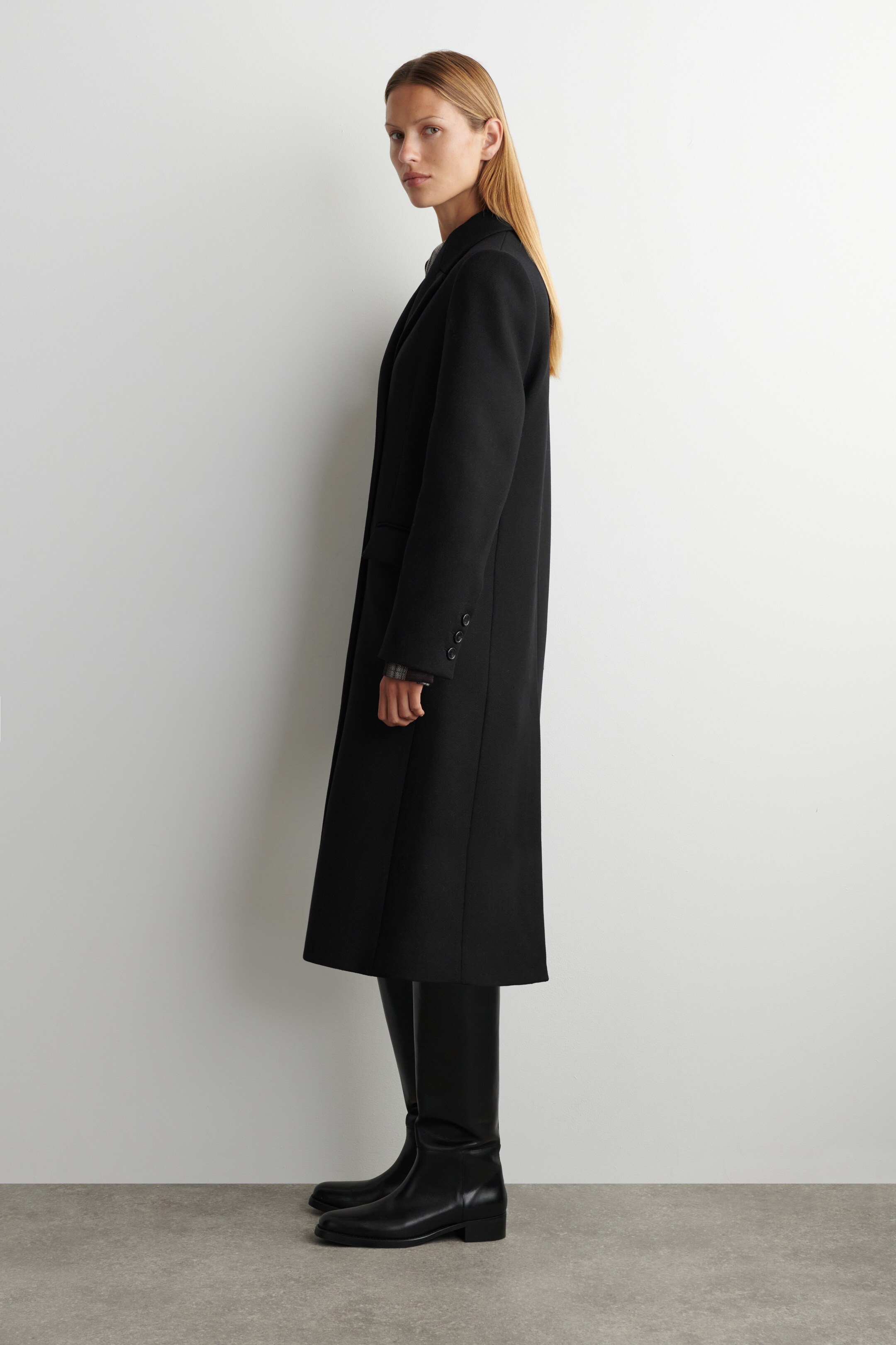 Ingrandisci l'immagine: Un lungo cappotto nero doppiopetto è indossato dalla modella, che si estende sotto le ginocchia e presenta tre bottoni su ciascun polsino. La modella è in piedi, abbinando il cappotto con stivali neri alti fino al ginocchio.