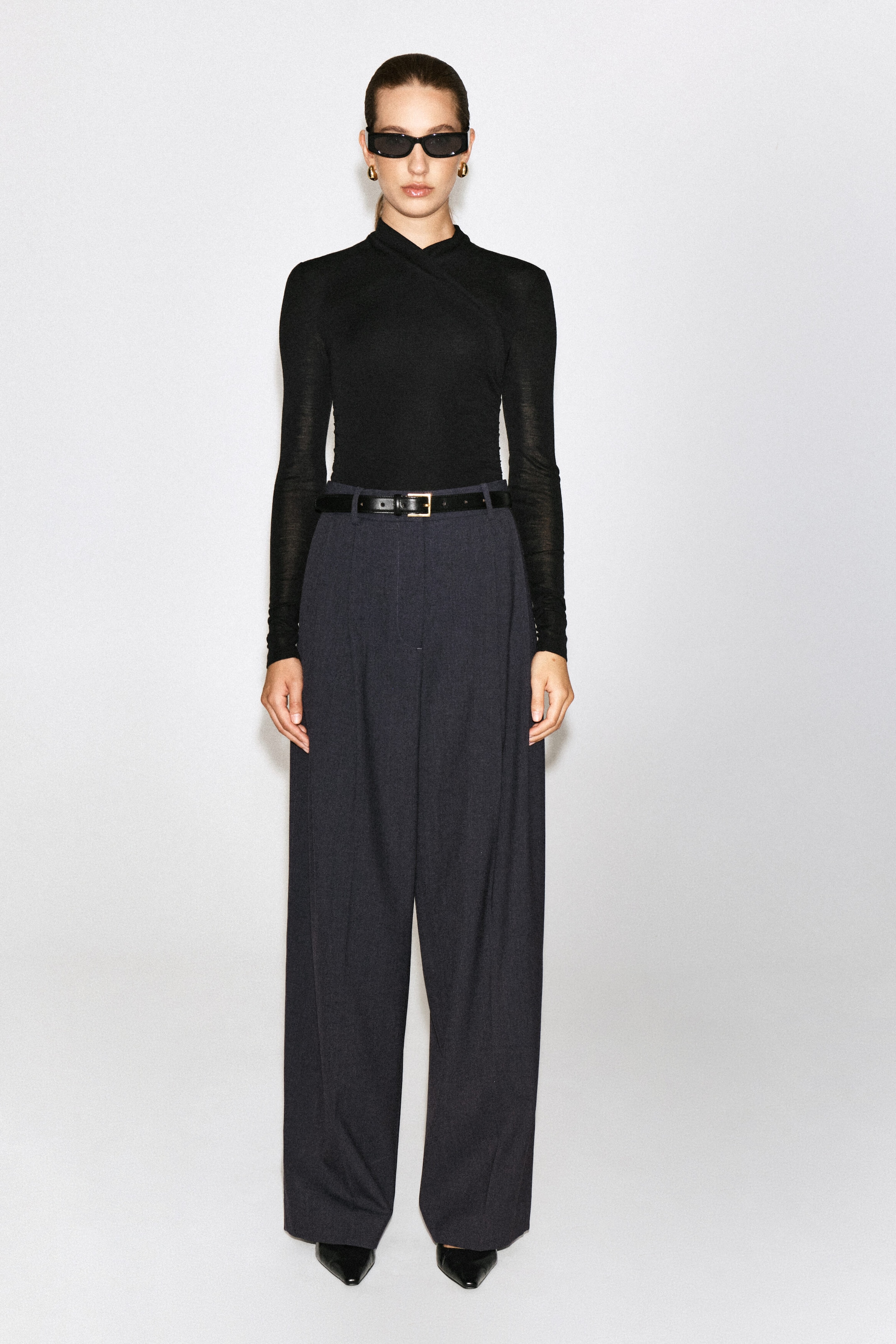 A black, slim-fit, long-sleeved top featuring a wrap-front shawl collar is being worn by the model. The fine-knit top is tucked into high-waisted, wide-leg dark trousers styled with a black belt. A black, slim-fit, long-sleeved top featuring a wrap-front shawl collar is being worn by the model. The fine-knit top is tucked into high-waisted, wide-leg dark trousers styled with a black belt.