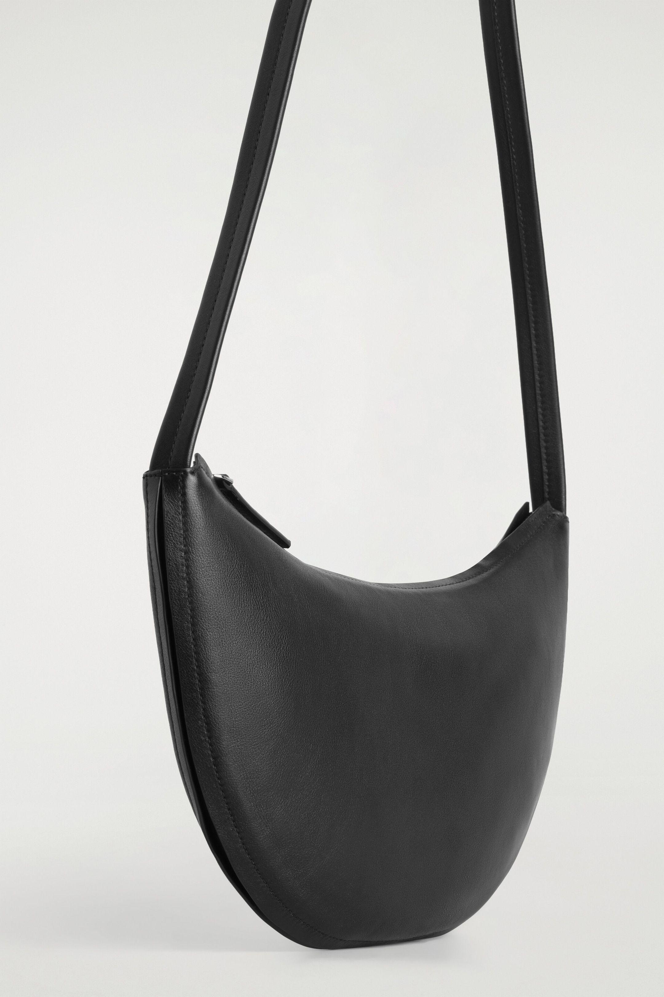 A black leather shoulder bag with a sculpted, crescent-shaped silhouette is hanging, showing off its smooth texture and structured form from a front three-quarter angle. The bag features a long shoulder strap and a zip closure along the top opening.