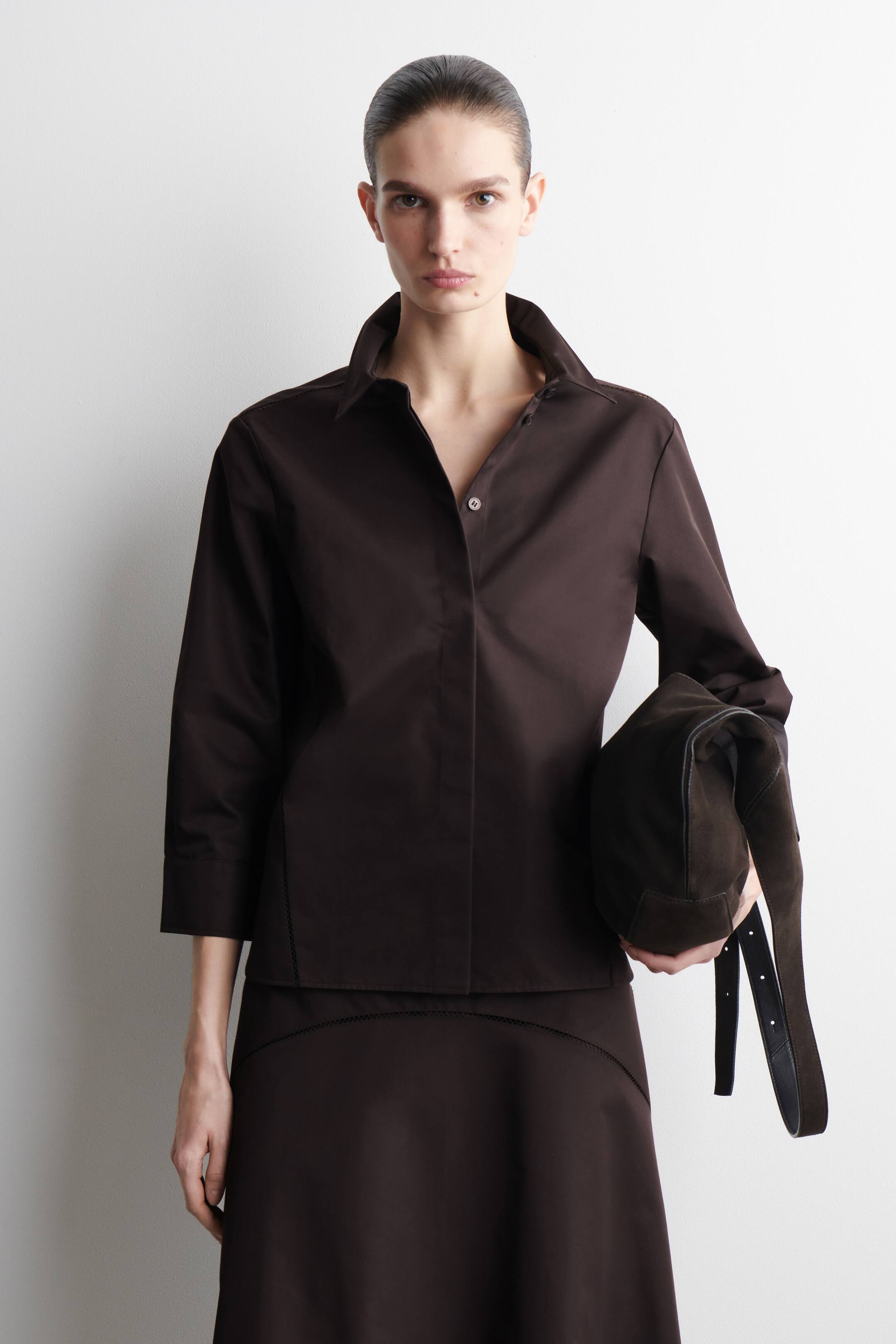 Προβολή μεγαλύτερης εικόνας: Ένα σκούρο καφέ πουκάμισο με κουμπιά, με όρθιο μυτερό γιακά και μανίκια τριών τετάρτων, φοριέται, διαθέτοντας ένα ανοιχτόχρωμο κουμπί και διακριτικές ραφές κατά μήκος μιας πλευρικής ραφής. Συνδυάζεται με μια αντίστοιχη σκούρα καφέ φούστα που έχει διακοσμητικές ραφές κατά μήκος μιας καμπύλης άνω ραφής, και μια σκούρα καφέ τσάντα κρατιέται.