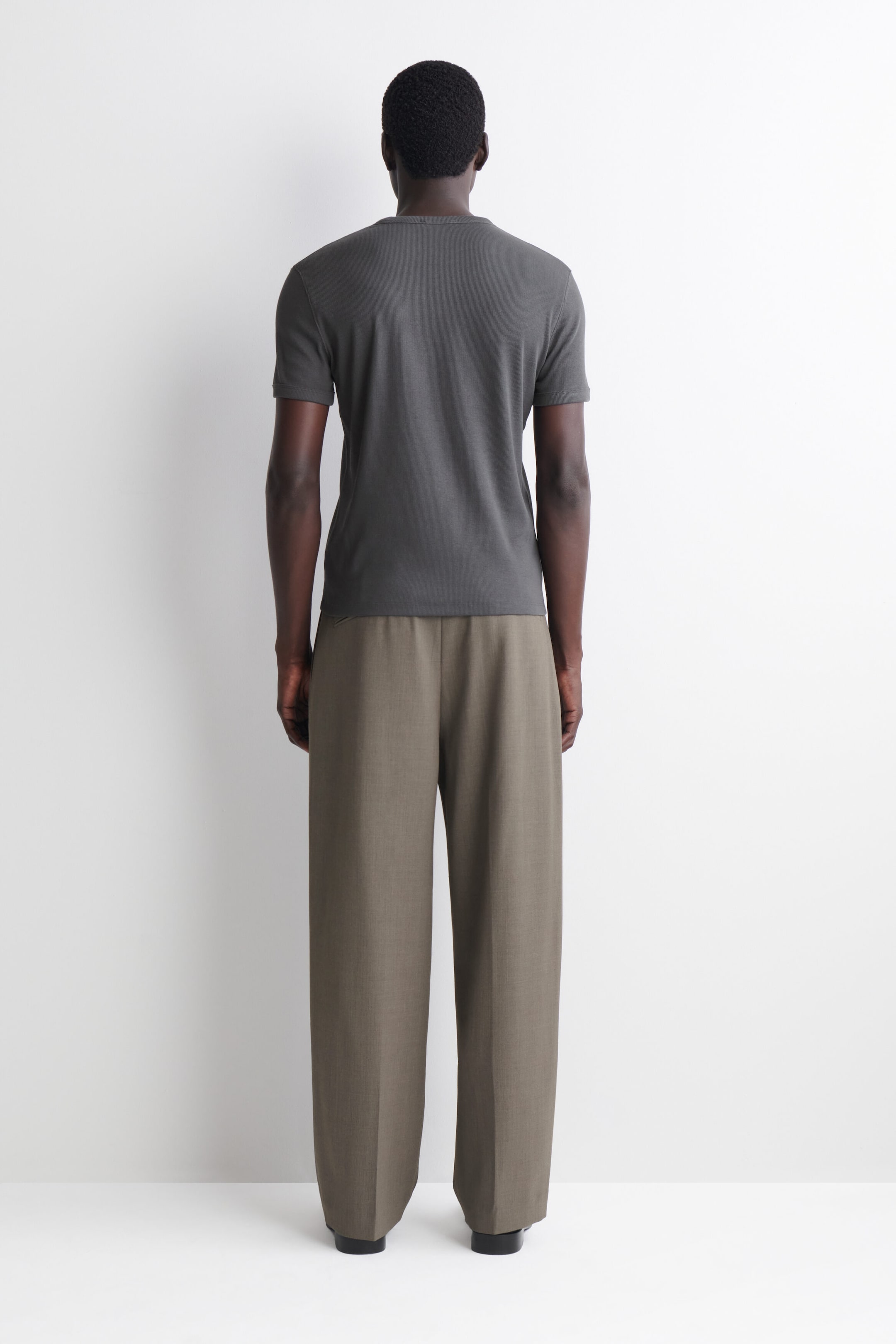 A dark grey slim-fit ribbed cotton T-shirt is shown from the back, highlighting its tailored silhouette, crew neck and short sleeves. The T-shirt sits neatly over the waistband of wide-leg trousers.