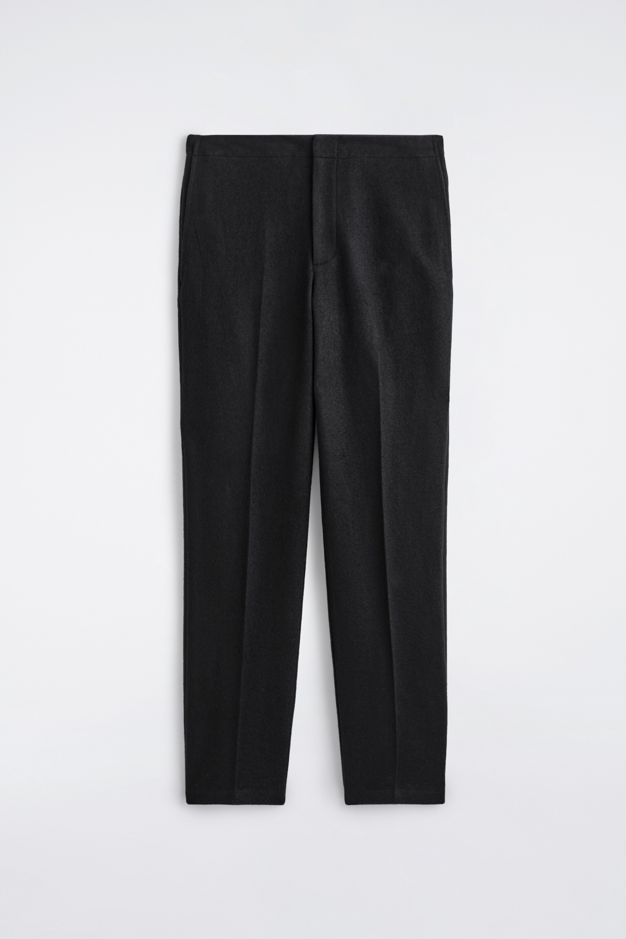 Agrandir l'image: Les PANTALONS EN LAINE BOUILLIE ÉLASTIQUÉS sont présentés de face, affichant une finition mate et sombre. Ils présentent une ceinture plate et ajustée avec une fermeture à glissière dissimulée. Des plis marqués descendent au centre de chaque jambe droite, se terminant par un ourlet net et non retourné.