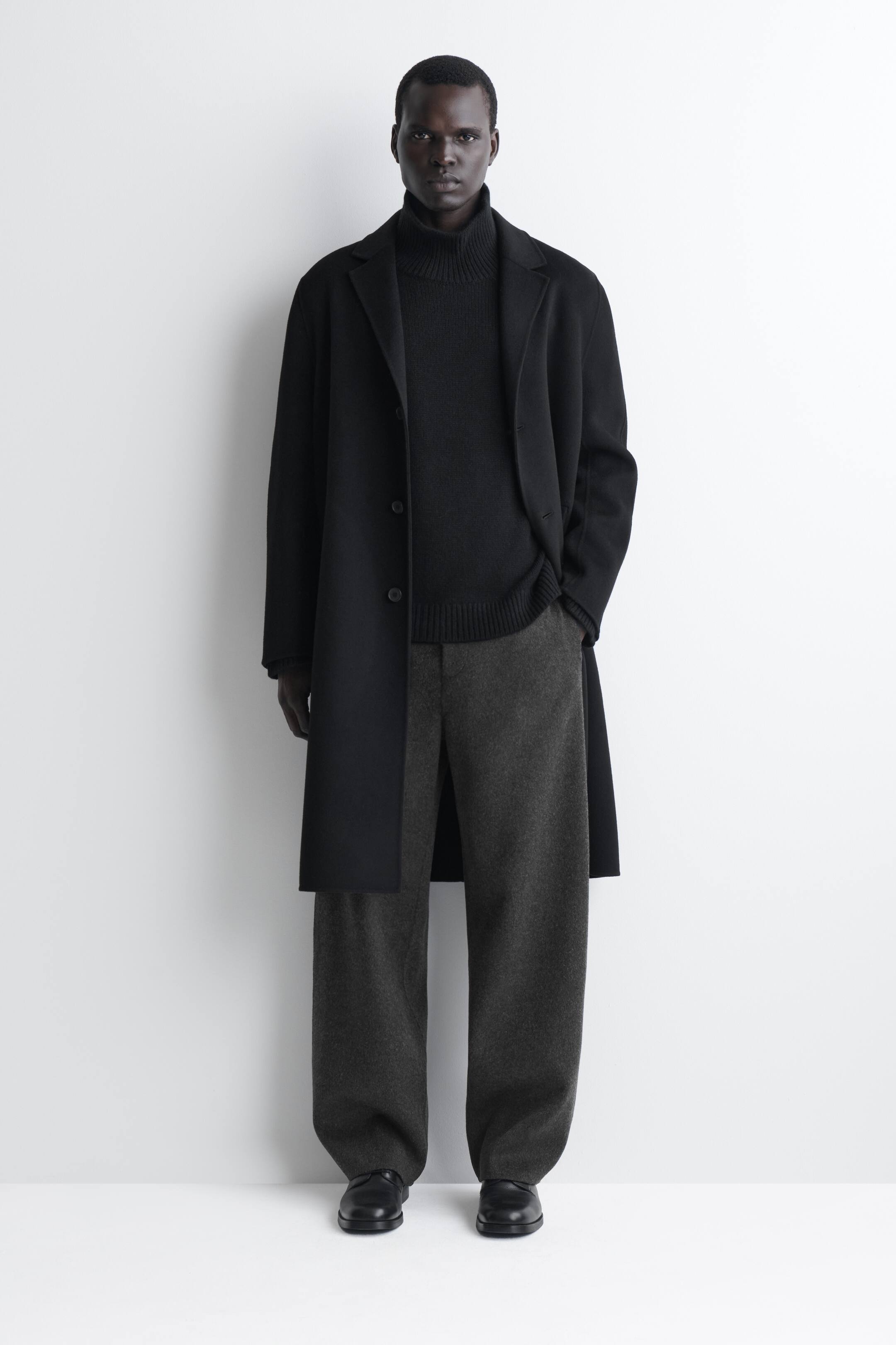 Agrandir l'image: Le pantalon ample à jambes larges en laine double face est présenté de face dans une teinte gris foncé, avec une coupe décontractée et un tombé large qui retombe légèrement sur des chaussures en cuir noir. Le pantalon est associé à un pull col roulé noir côtelé et à un long manteau noir ouvert, mettant en valeur sa silhouette décontractée.