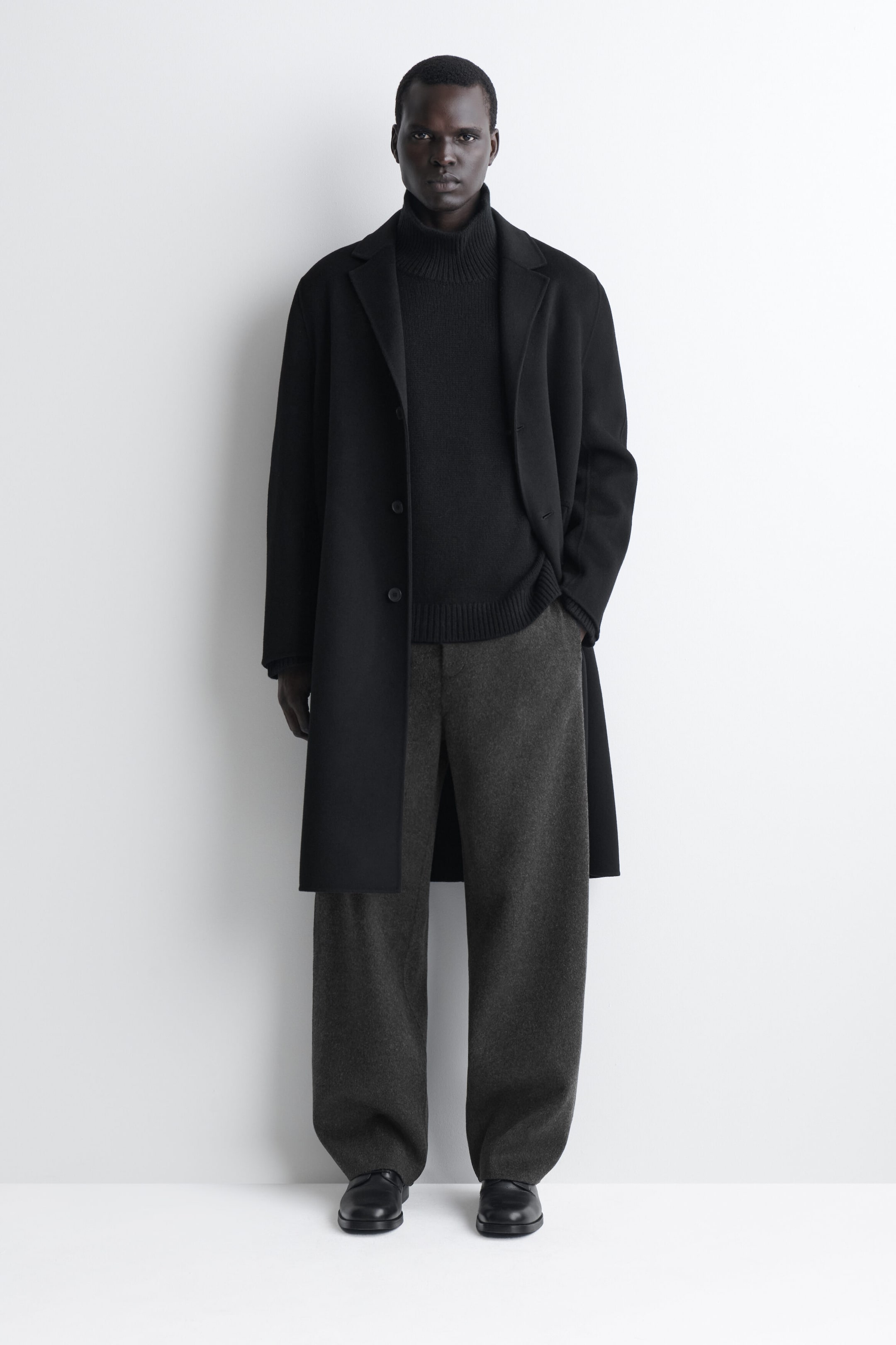 Die doppelseitig gewebte Wollhose im Relaxed Fit mit weitem Bein ist von vorne in Dunkelgrau zu sehen und besticht durch ihren lockeren, weit fallenden Schnitt, der leicht über schwarze Lederschuhe fällt. Die Hose wird mit einem schwarzen, gerippten Rollkragenpullover und einem offenen, langen schwarzen Mantel kombiniert, wodurch ihre entspannte Silhouette betont wird. Die doppelseitig gewebte Wollhose im Relaxed Fit mit weitem Bein ist von vorne in Dunkelgrau zu sehen und besticht durch ihren lockeren, weit fallenden Schnitt, der leicht über schwarze Lederschuhe fällt. Die Hose wird mit einem schwarzen, gerippten Rollkragenpullover und einem offenen, langen schwarzen Mantel kombiniert, wodurch ihre entspannte Silhouette betont wird.