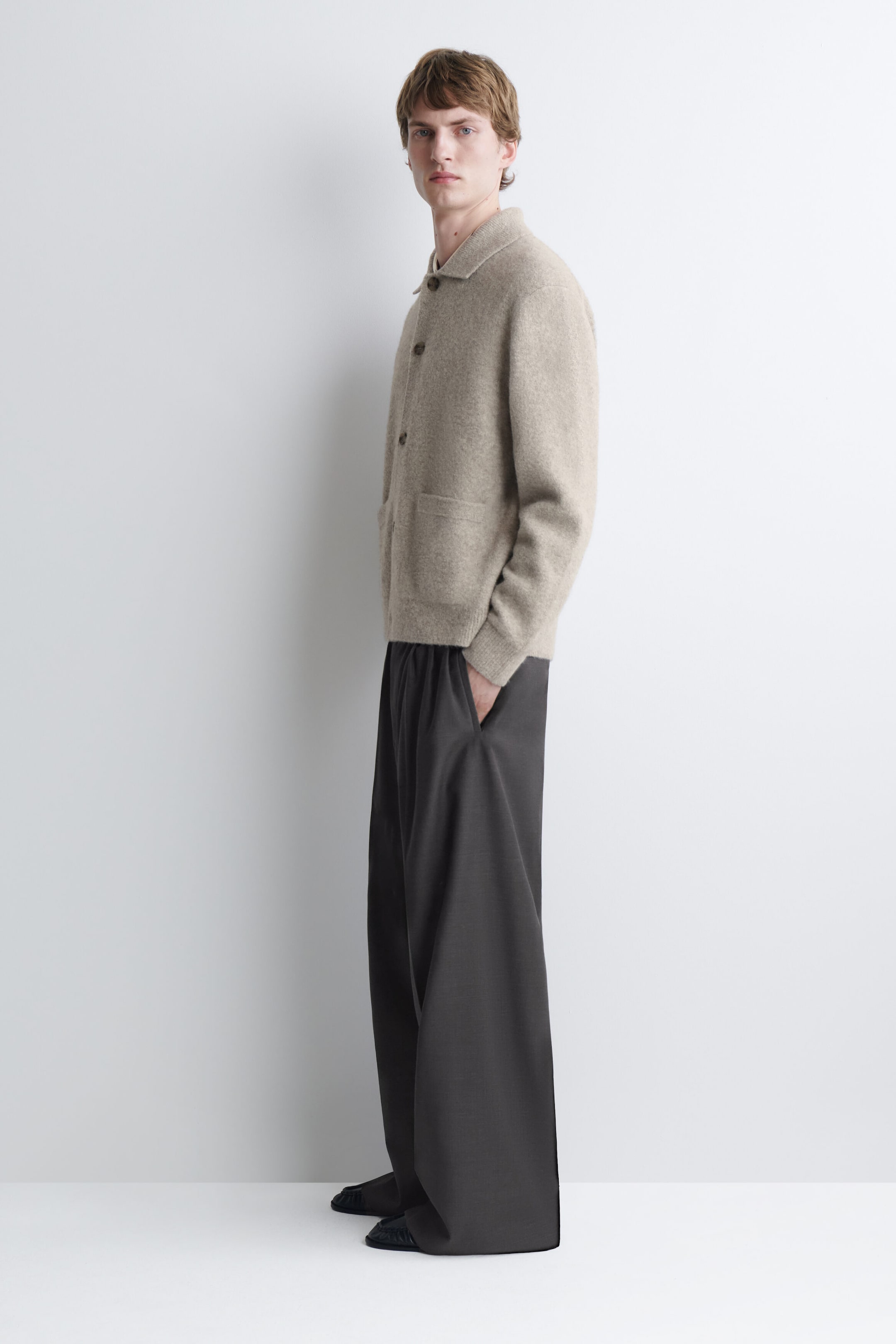 A beige melange knit cardigan featuring a classic collar, button closure, and patch pockets is worn by the model. The cardigan is styled with dark, wide-leg trousers and dark shoes. A beige melange knit cardigan featuring a classic collar, button closure, and patch pockets is worn by the model. The cardigan is styled with dark, wide-leg trousers and dark shoes.