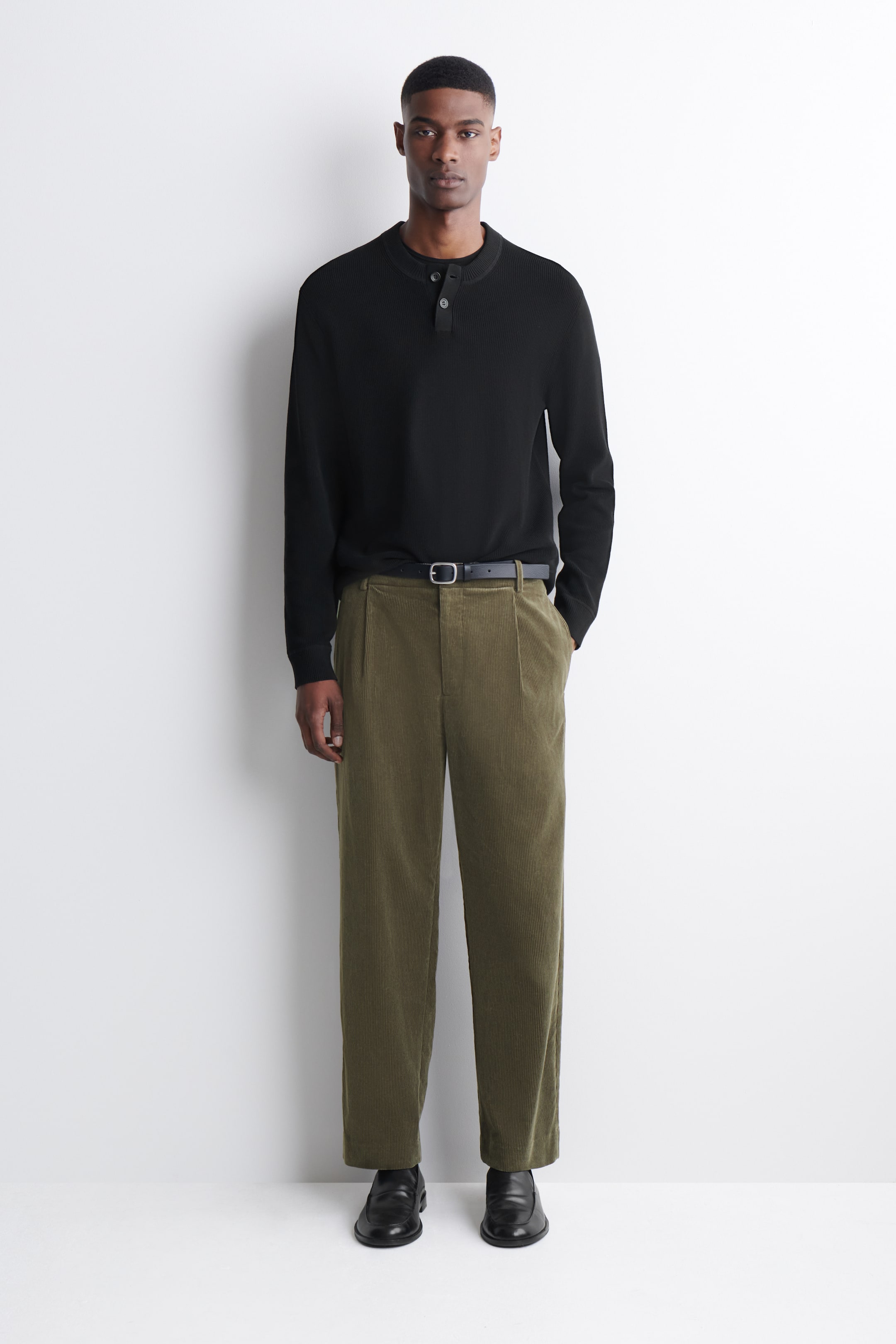 Grösseres Bild ansehen: Ein schwarzes Langarm-Henley-Shirt mit einer Zwei-Knopf-Leiste und einer strukturierten Strickoptik wird von vorne präsentiert, wobei ein schwarzes Rundhals-Unterhemd darunter sichtbar ist. Es wird mit olivgrünen, plissierten Cordhosen kombiniert, die durch einen schwarzen Gürtel zusammengehalten werden, und schwarzen Slippern.