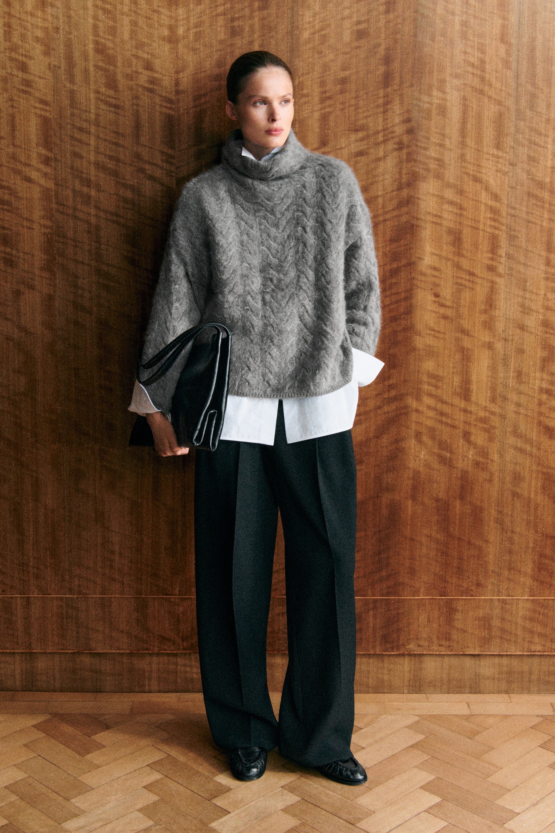 The grey cable-knit brushed-cashmere funnel-neck jumper is shown from the front, featuring a relaxed, oversized silhouette and a textured vertical cable pattern. It is layered over a white collared shirt that extends past the ribbed hem and cuffs, emphasising the jumper’s slouchy collar and loose fit. The grey cable-knit brushed-cashmere funnel-neck jumper is shown from the front, featuring a relaxed, oversized silhouette and a textured vertical cable pattern. It is layered over a white collared shirt that extends past the ribbed hem and cuffs, emphasising the jumper’s slouchy collar and loose fit.