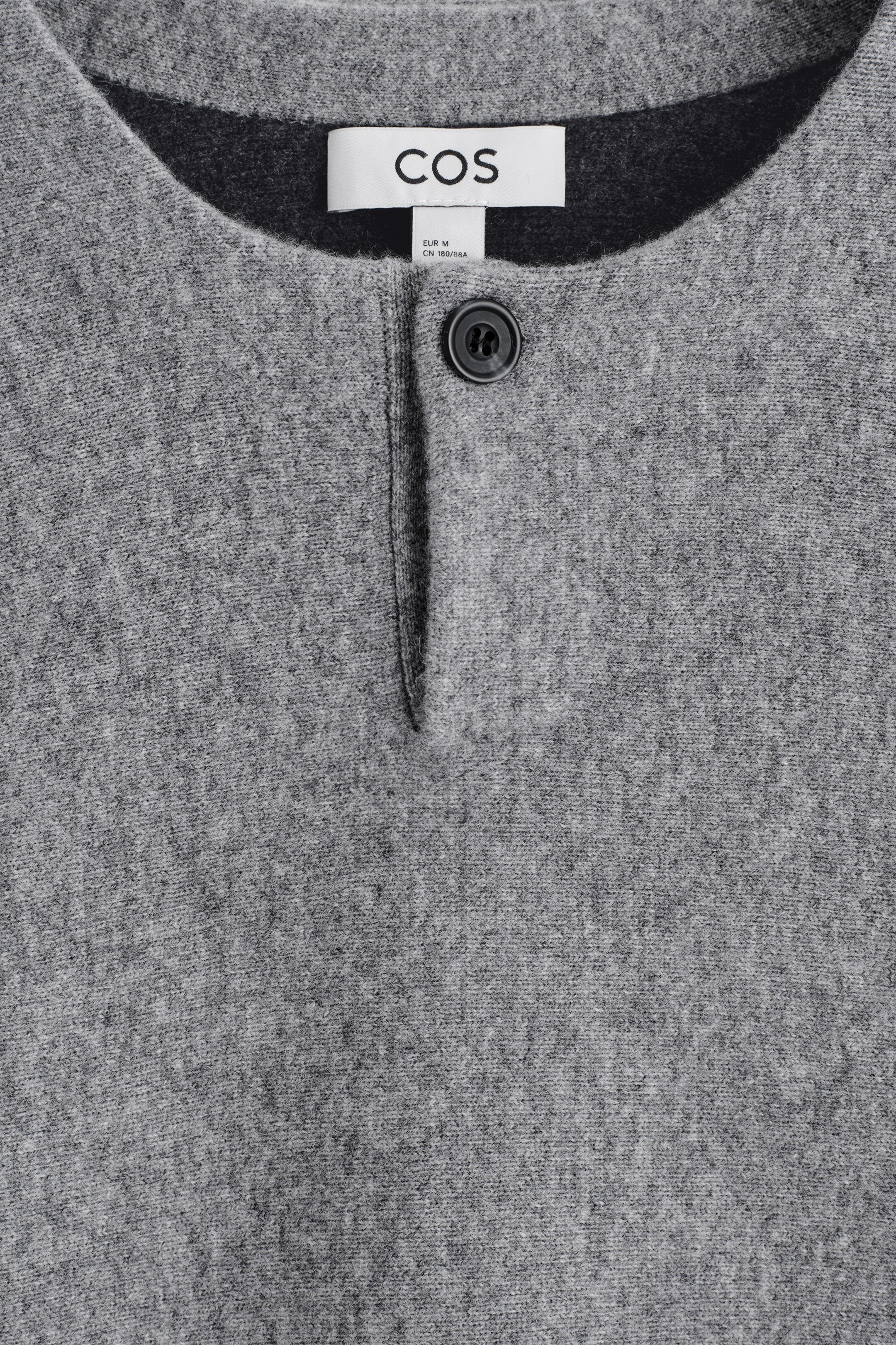 Agrandir l'image: Un pull en tricot gris clair est montré de face, avec un col rond et une patte de boutonnage partielle avec un bouton foncé à quatre trous visible. Une étiquette en tissu blanc avec "COS" en texte noir est visible à l'arrière du cou.