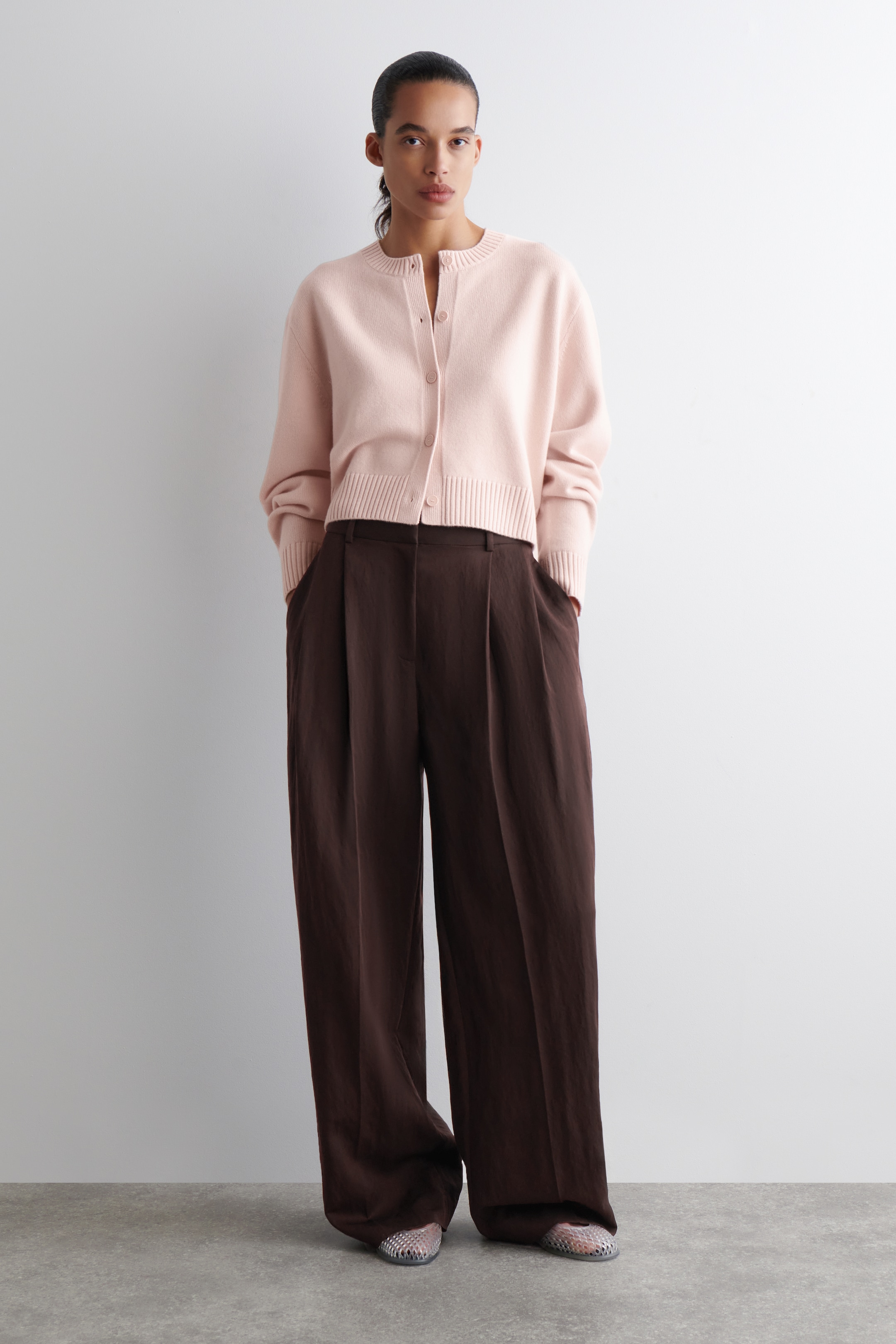 Agrandir l'image: Un cardigan court rose clair, avec un col rond, une fermeture à boutons sur le devant, et des poignets et un ourlet côtelés, est porté de face. Il est assorti à un pantalon large marron foncé, qui présente des plis à l'avant et des poches latérales visibles, avec des ballerines argentées texturées qui dépassent sous les ourlets.
