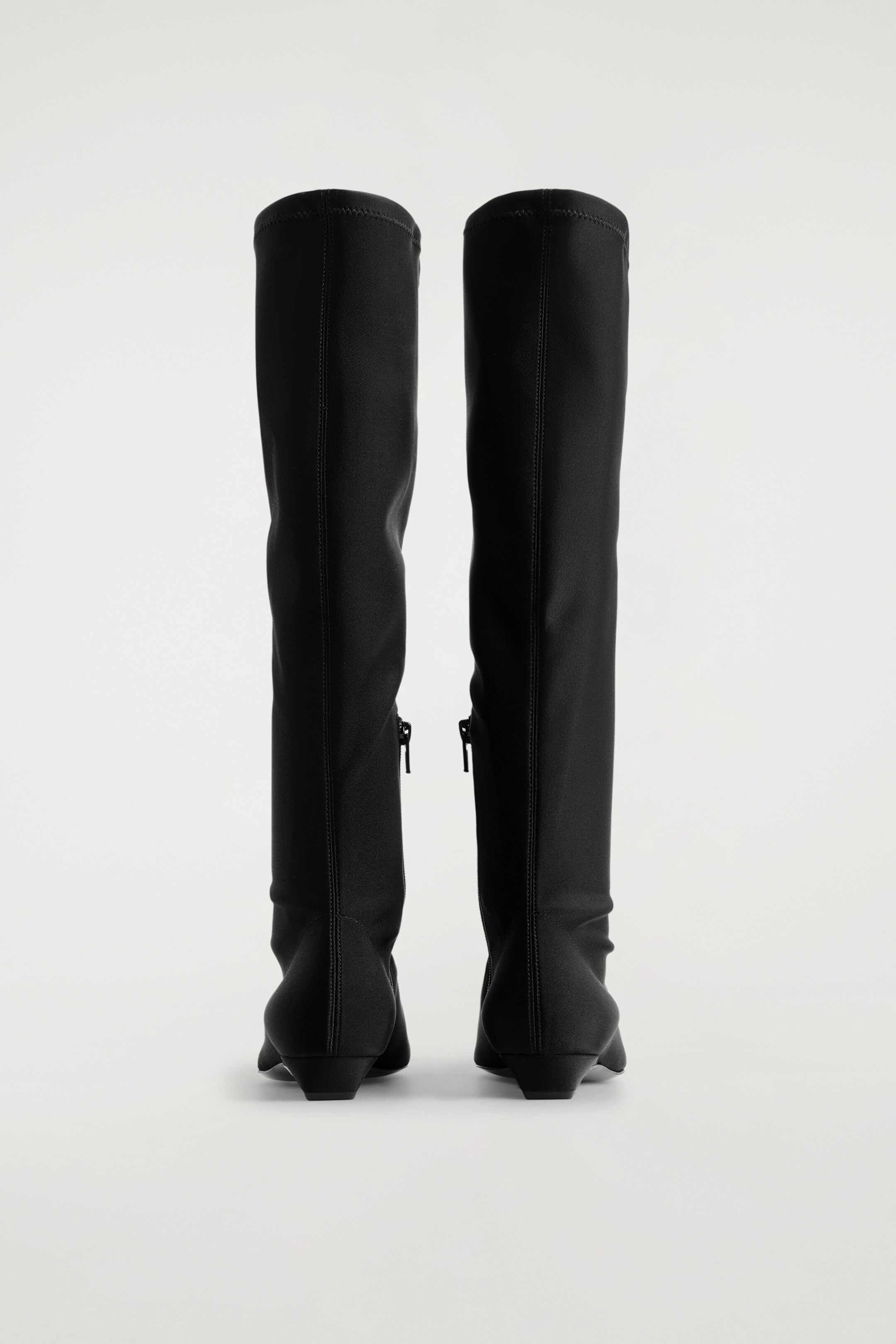Größeres Bild ansehen: RÜCKANSICHT von schwarzen, spitzen Scuba-Kniestrümpfstiefeln mit einer glatten, dehnbaren Textur. Jeder Stiefel zeigt eine vertikale Naht, die den Rücken seines Schafts hinauf verläuft, einen sichtbaren Reißverschluss auf der Innenseite, der sich vom Knöchel aus erstreckt, und steht auf einem niedrigen, passenden Blockabsatz.