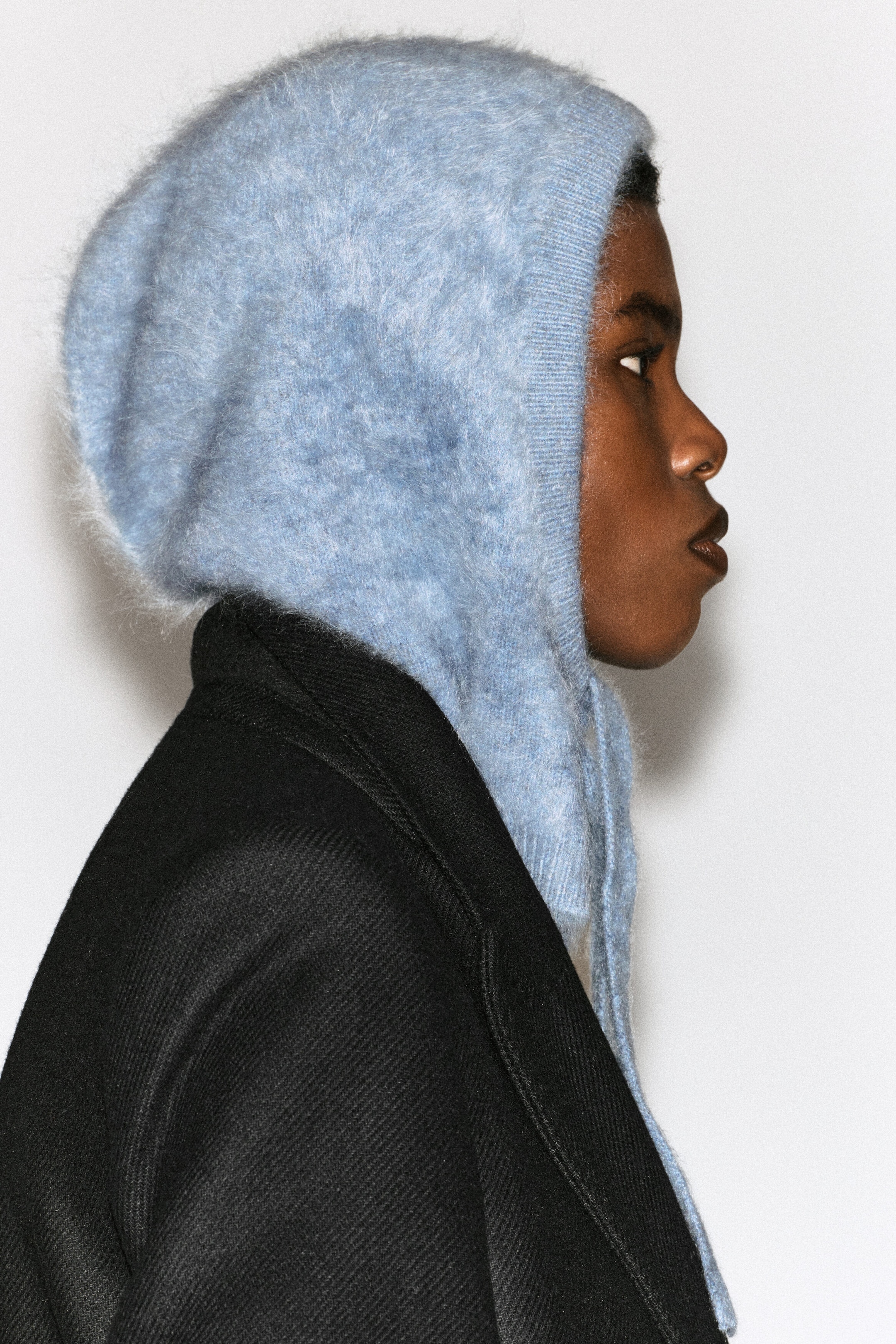 A light blue knitted hood with a fuzzy, brushed texture is being worn by the model in profile. The hood features a ribbed trim around the face and hanging ties, styled over a black textured coat.