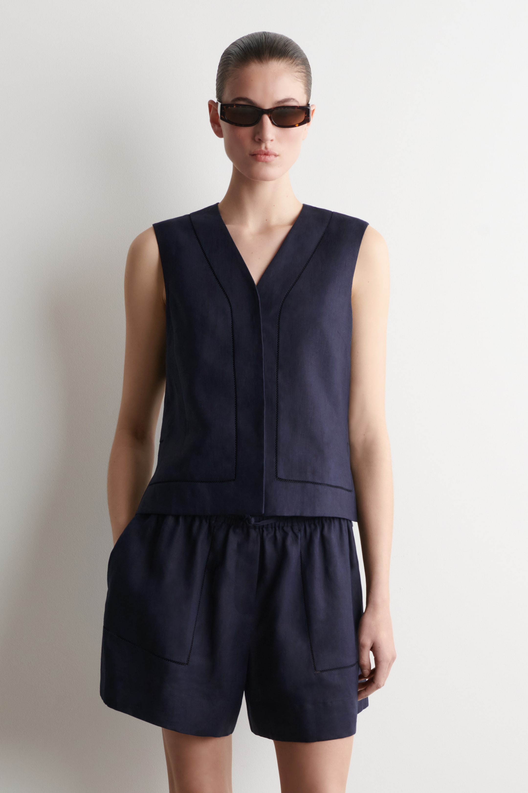 Grotere afbeelding bekijken: Een marineblauw linnen gilet met een V-hals, knoopsluiting aan de voorkant en geborduurde details langs de randen en voorpanelen wordt gedragen door het model. Het model staat met de handen in de zakken van bijpassende marineblauwe linnen shorts, die ook voorzien zijn van geborduurde details.