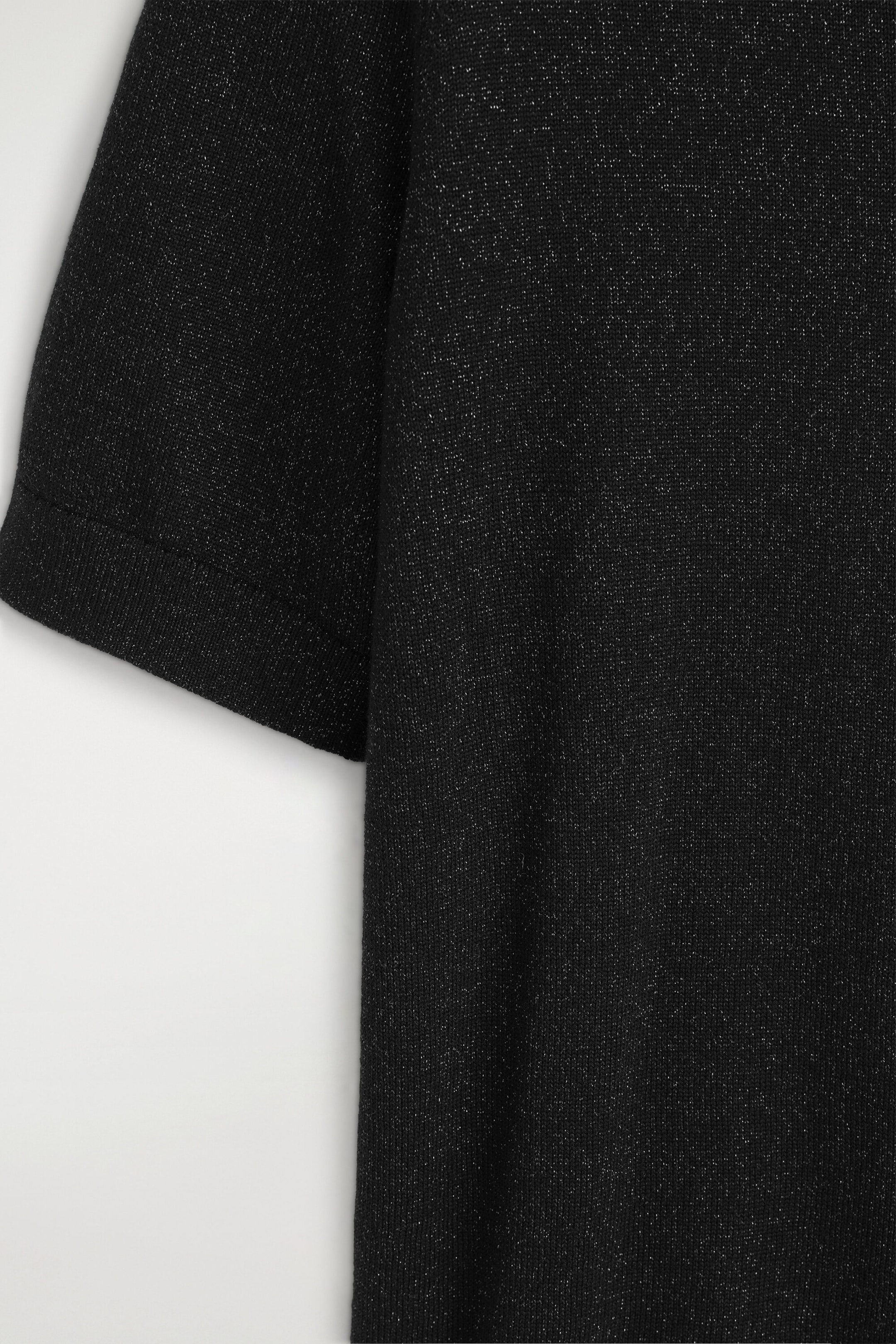 A close-up view showing the fine-knit black fabric of a T-shirt featuring subtle metallic flecks that create a shimmering effect. The image highlights the texture of the knit and a short sleeve ending with a ribbed cuff. A close-up view showing the fine-knit black fabric of a T-shirt featuring subtle metallic flecks that create a shimmering effect. The image highlights the texture of the knit and a short sleeve ending with a ribbed cuff.