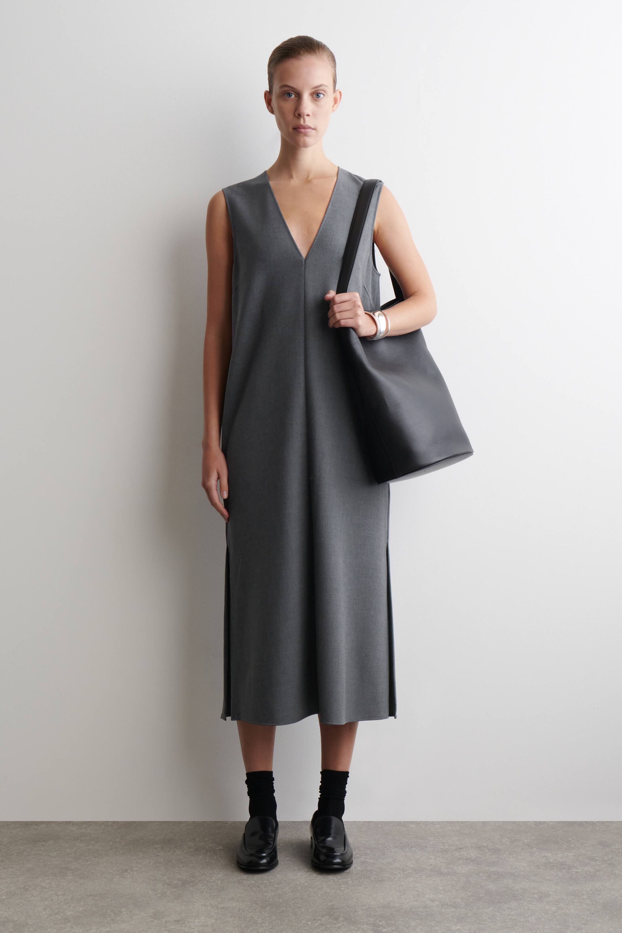 הצגת תמונה גדולה יותר: שמלת מידי אפורה ללא שרוולים עם מחשוף וי מוצגת מזווית קדמית, ויש לה גזרה נינוחה ופתחים בצדדים. תיק טוט מעור שחור מחוספס נישא על הכתף הימנית.