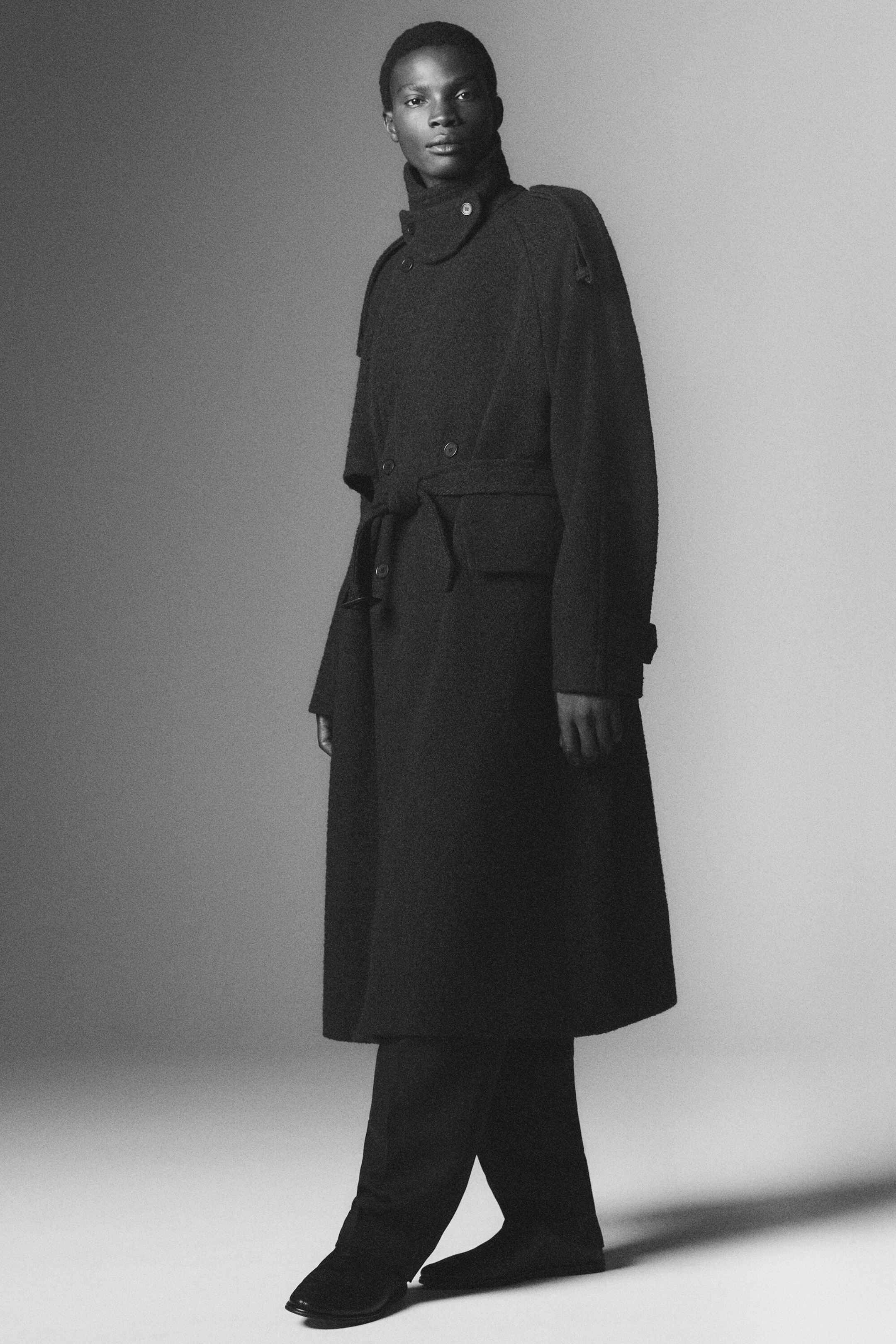 Ein langer, dunkler, zweireihiger Trenchcoat ist aus einer Dreiviertelansicht zu sehen. Er verfügt über einen hohen Kragen mit geknöpftem Kinnriemen, Schulterepauletten und einen selbst zu bindenden Gürtel in der Taille. Der Mantel fällt in einer legeren Passform bis unter das Knie und ist mit passenden dunklen Hosen und Schuhen kombiniert. Ein langer, dunkler, zweireihiger Trenchcoat ist aus einer Dreiviertelansicht zu sehen. Er verfügt über einen hohen Kragen mit geknöpftem Kinnriemen, Schulterepauletten und einen selbst zu bindenden Gürtel in der Taille. Der Mantel fällt in einer legeren Passform bis unter das Knie und ist mit passenden dunklen Hosen und Schuhen kombiniert.