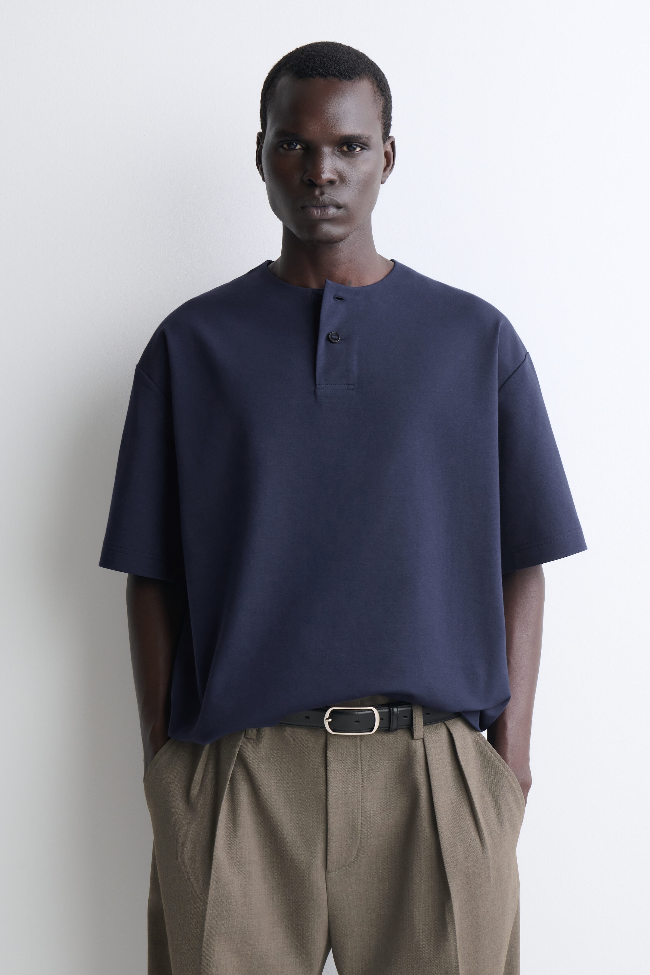 Un t-shirt Henley oversize en jersey de coton bleu marine est présenté de face, avec une coupe décontractée, des épaules tombantes et de larges manches courtes. Le t-shirt affiche une courte patte de boutonnage avec deux boutons au col rond et est porté rentré de façon souple dans un pantalon plissé avec une ceinture pour mettre en valeur sa structure drapée.