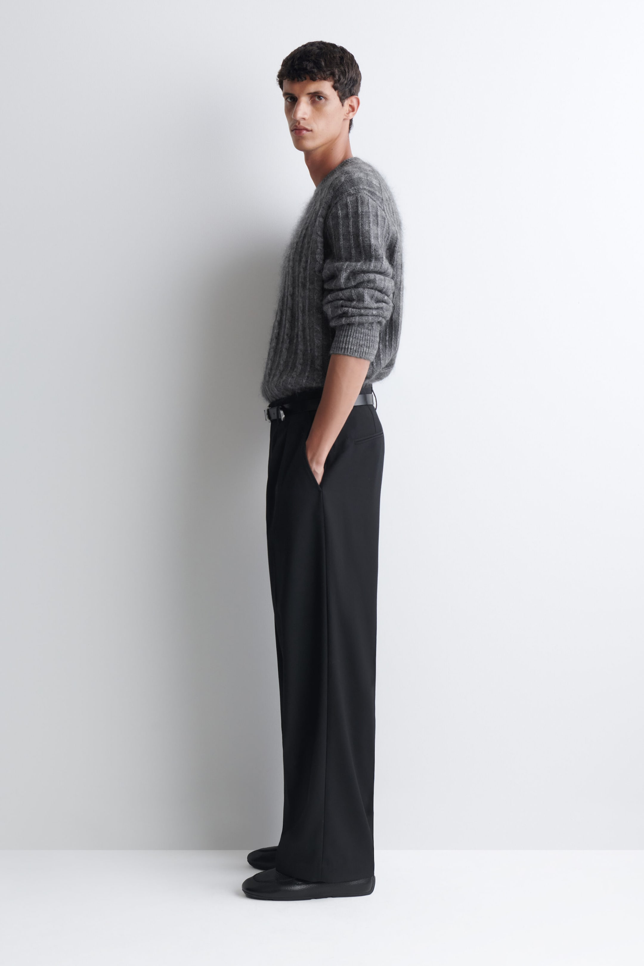 A grey, textured ribbed-knit sweater is worn by the model and tucked into black wide-leg trousers. The sweater features long sleeves pushed up to the forearms, and the trousers are accessorized with a black belt, with the model resting one hand in a pocket. A grey, textured ribbed-knit sweater is worn by the model and tucked into black wide-leg trousers. The sweater features long sleeves pushed up to the forearms, and the trousers are accessorized with a black belt, with the model resting one hand in a pocket.