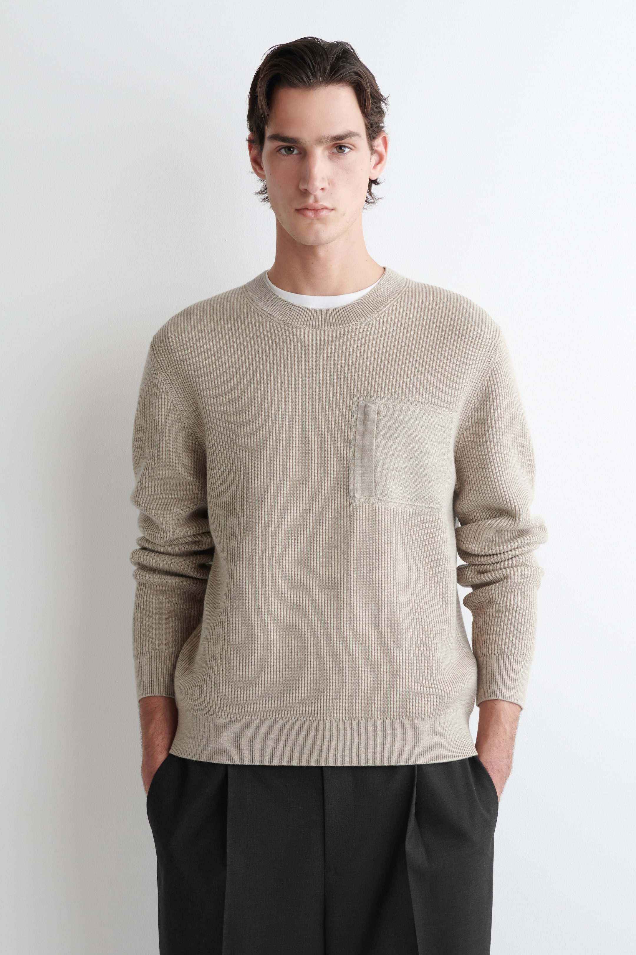 Agrandir l'image: Un pull en maille côtelée beige présente son devant, avec un col rond, des manches longues avec des poignets côtelés et un ourlet côtelé. Une poche plaquée rectangulaire avec une texture plus lisse est située sur la poitrine gauche, et un t-shirt blanc à col rond est visible sous l'encolure.