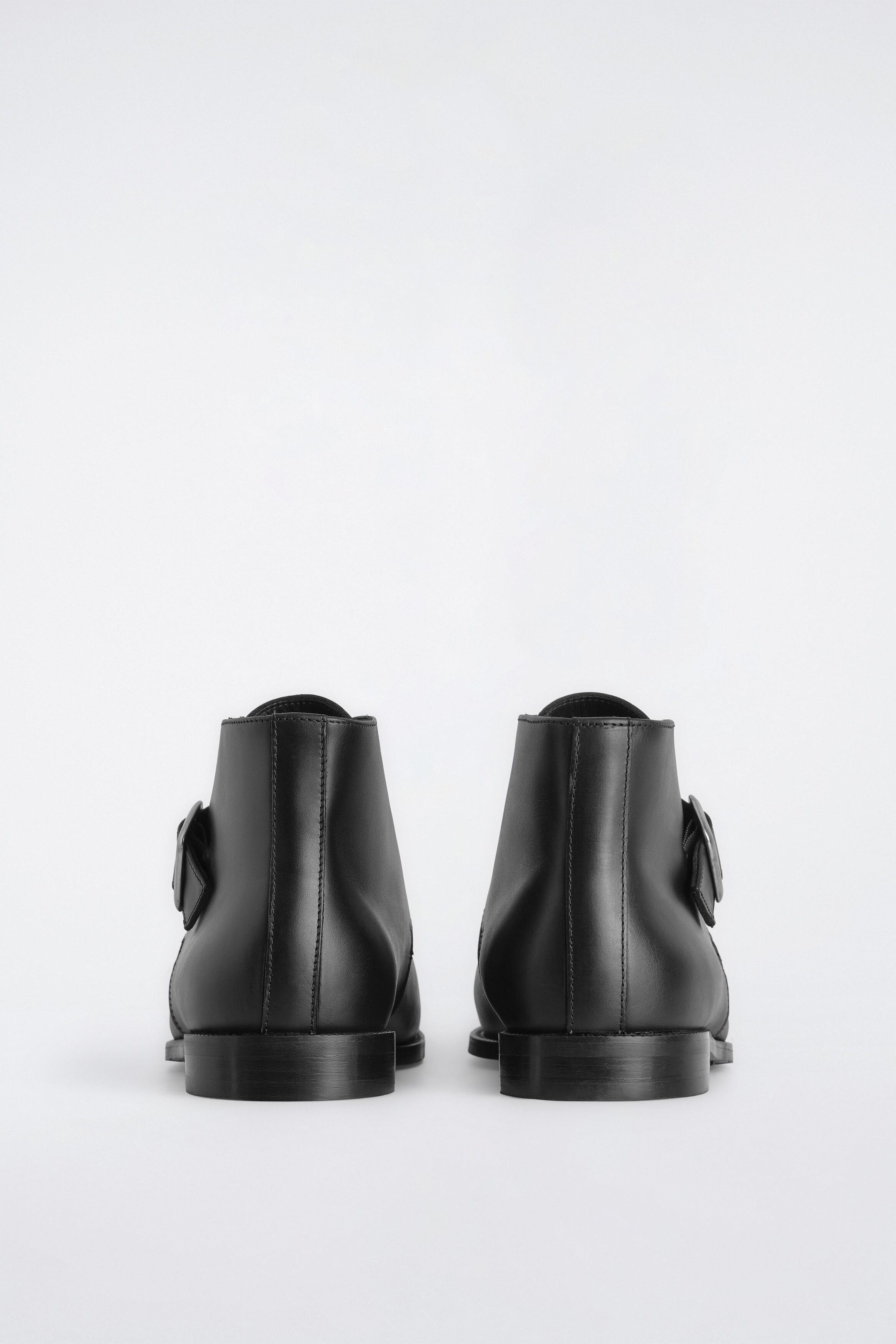 Ein Paar schwarze Leder-Monkstiefel steht nebeneinander und wird von hinten gezeigt, um die glatte Lederverarbeitung, die vertikalen Nähte an der Ferse und die niedrigen Blockabsätze hervorzuheben. Die mit Schnallen versehenen Knöchelriemen sind an den Außenseiten jedes Stiefels sichtbar.