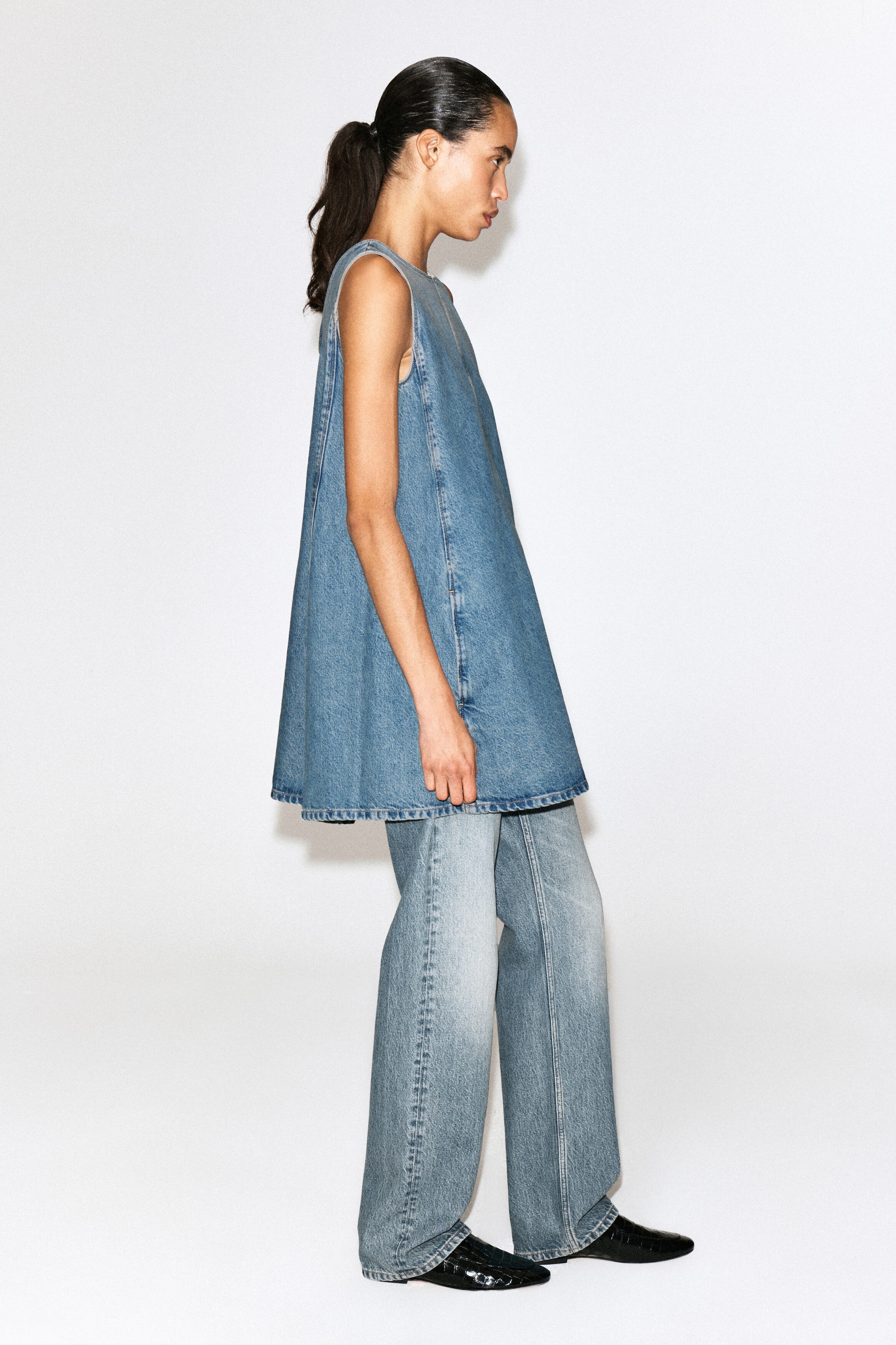 Un mini abito in denim senza maniche, in lavaggio blu medio, è mostrato in una vista di profilo rivolto a destra, con silhouette svasata a trapezio e cuciture impunturate strutturate. L’abito è indossato sopra jeans in denim coordinati a gamba ampia, mettendo in risalto il volume e la versatilità di styling.