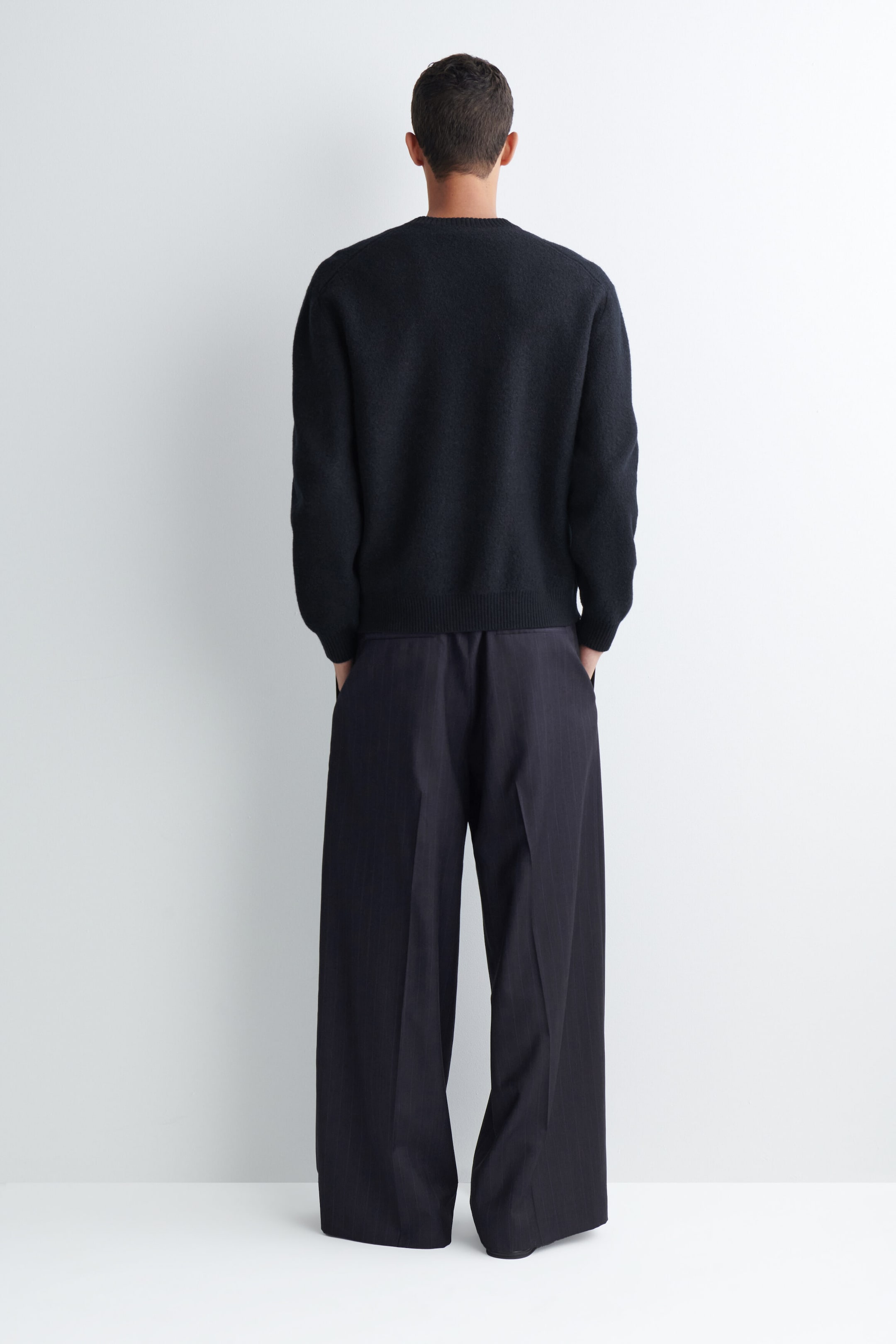 Pantalon ample à jambes larges en tissu gris foncé, avec plis, porté par le mannequin et présenté de dos, les mains placées dans les poches latérales. Le pantalon présente un pli marqué au centre de каждой jambe et est agencé avec un chandail noir à col rond à manches longues. Pantalon ample à jambes larges en tissu gris foncé, avec plis, porté par le mannequin et présenté de dos, les mains placées dans les poches latérales. Le pantalon présente un pli marqué au centre de каждой jambe et est agencé avec un chandail noir à col rond à manches longues.