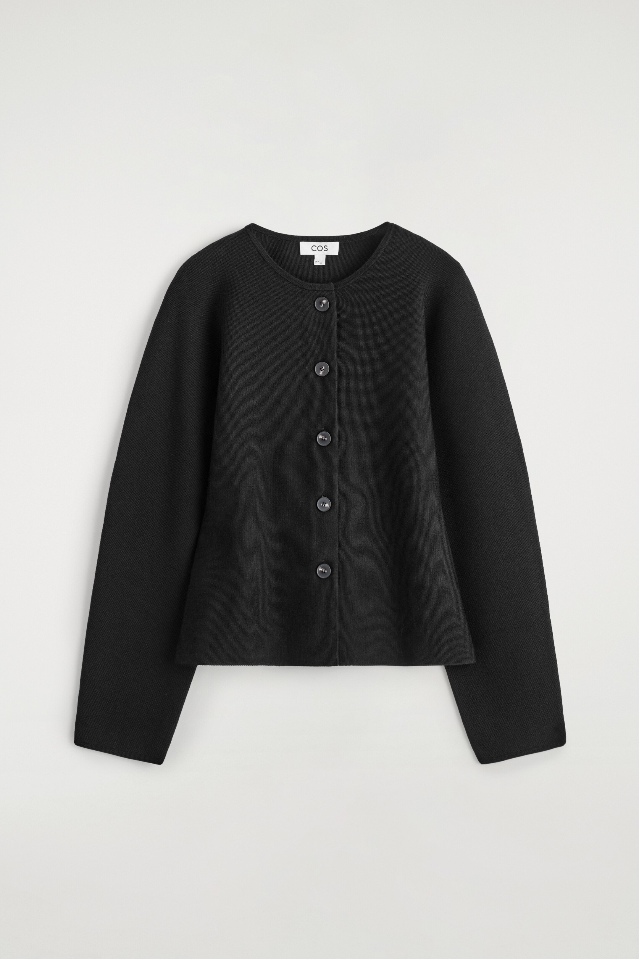 Größeres Bild ansehen: Ein schwarzer Cardigan wird von vorne gezeigt, mit einem runden Halsausschnitt und einem vollständigen Knopfverschluss mit fünf dunklen, runden Knöpfen. Er zeigt lange Ärmel und einen strukturierten, leicht ausgestellten Saum, der eine skulpturale Silhouette schafft.