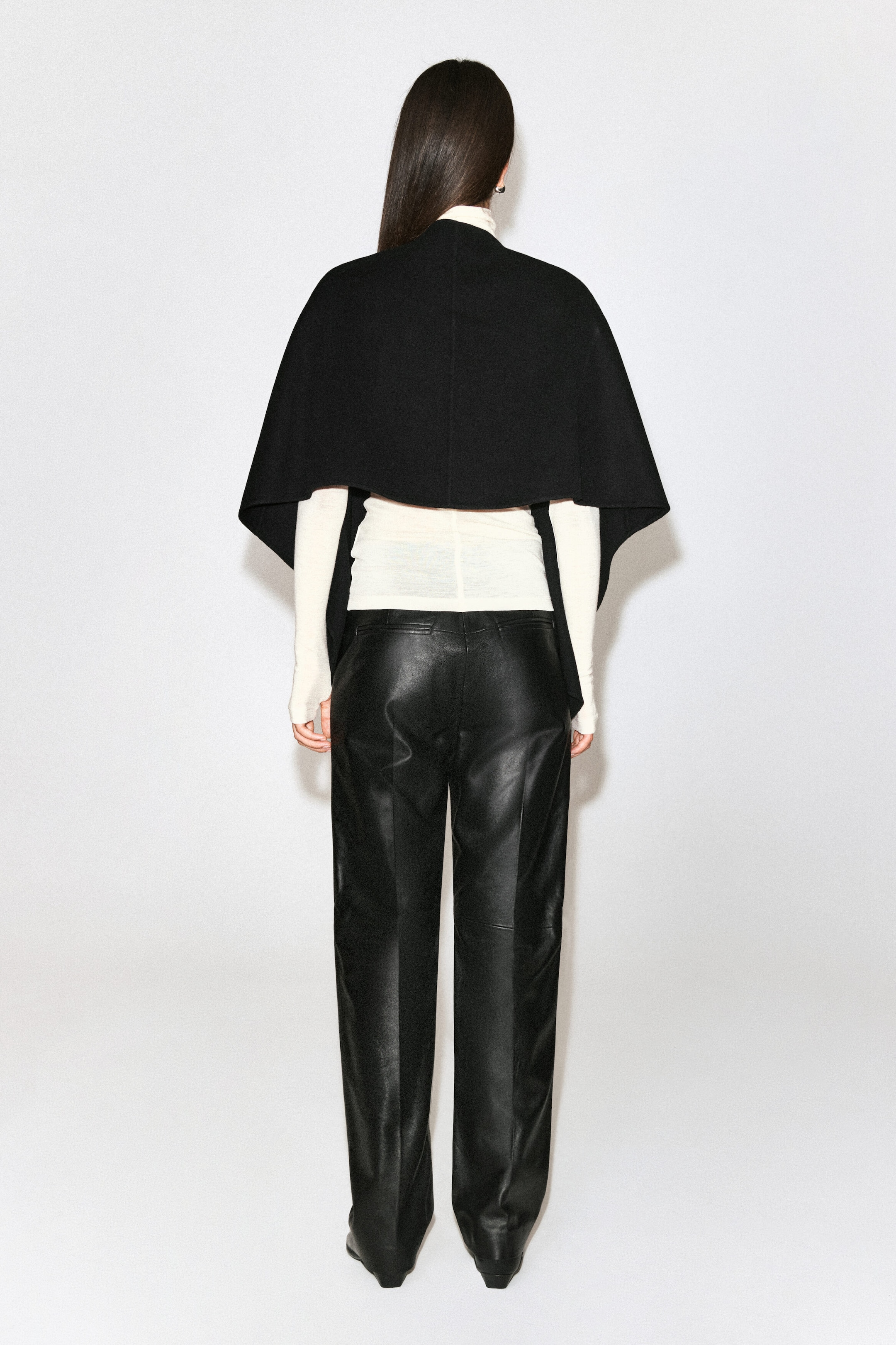 A black double-faced wool-blend cape is being worn by the model, draping over the shoulders to show a cropped back hem. The cape is layered over a cream long-sleeved roll-neck top and straight-leg black leather trousers. A black double-faced wool-blend cape is being worn by the model, draping over the shoulders to show a cropped back hem. The cape is layered over a cream long-sleeved roll-neck top and straight-leg black leather trousers.