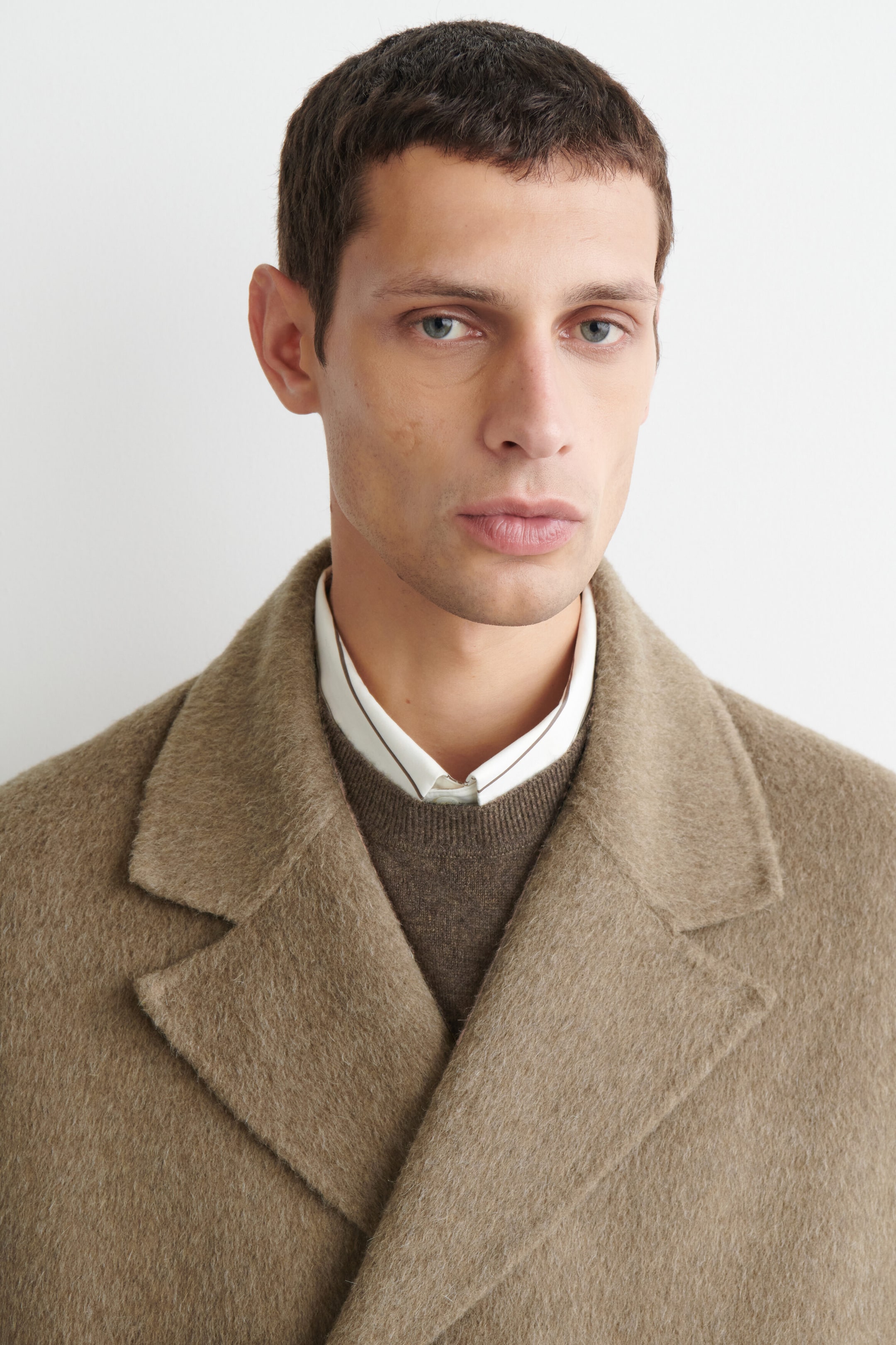 The double-faced wool-blend long coat is shown in a close-up shot focusing on the top half, highlighting its wide notch lapels and textured light brown fabric. The coat is layered over a brown crewneck sweater and a white collared shirt, emphasizing its structured shoulder design and collar fit.
