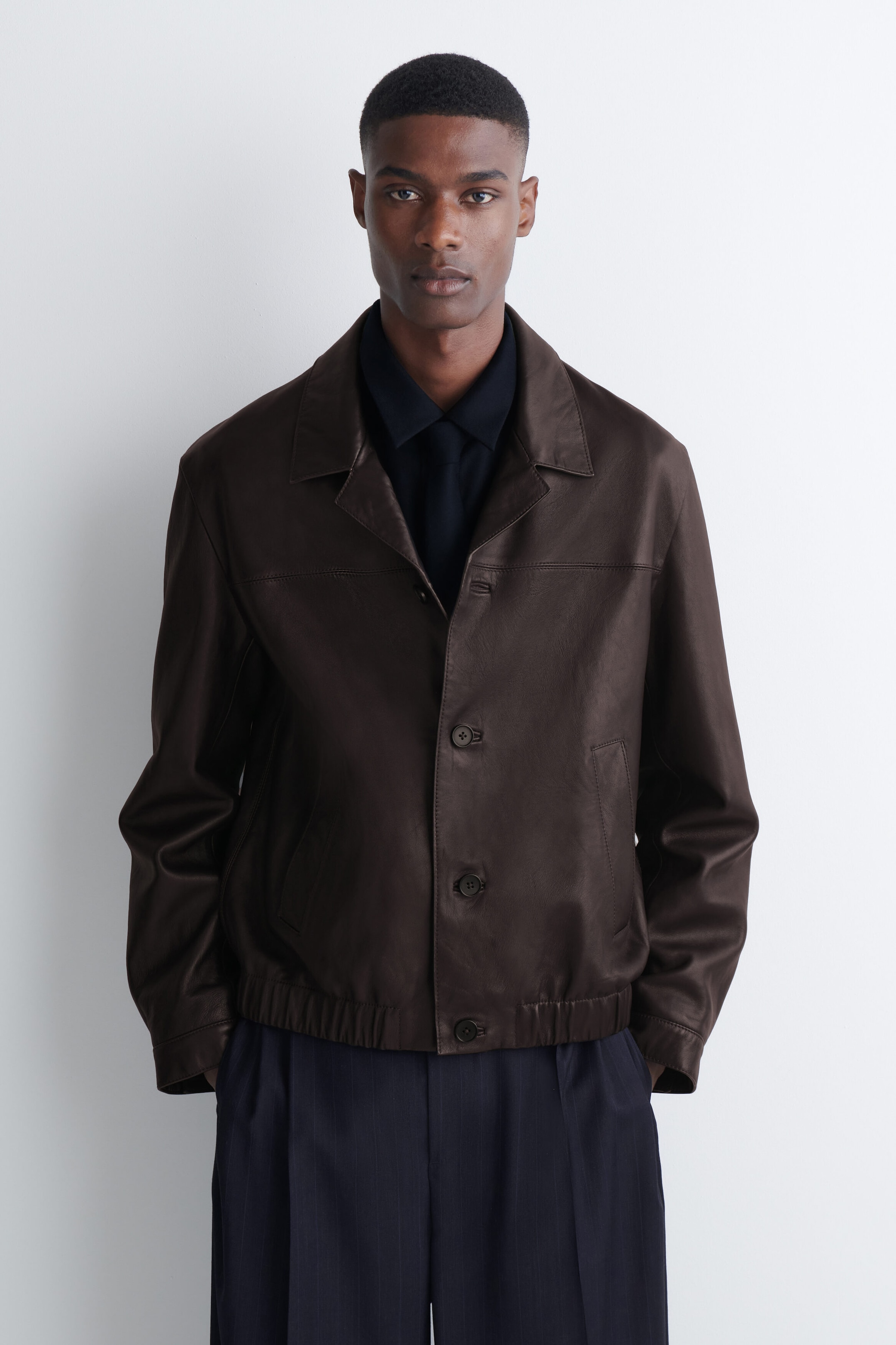 The model is wearing a dark wool flannel tie, styled with a knotted finish beneath a matching dark collared shirt. The tie is layered under a dark brown leather jacket to complete the outfit. The model is wearing a dark wool flannel tie, styled with a knotted finish beneath a matching dark collared shirt. The tie is layered under a dark brown leather jacket to complete the outfit.