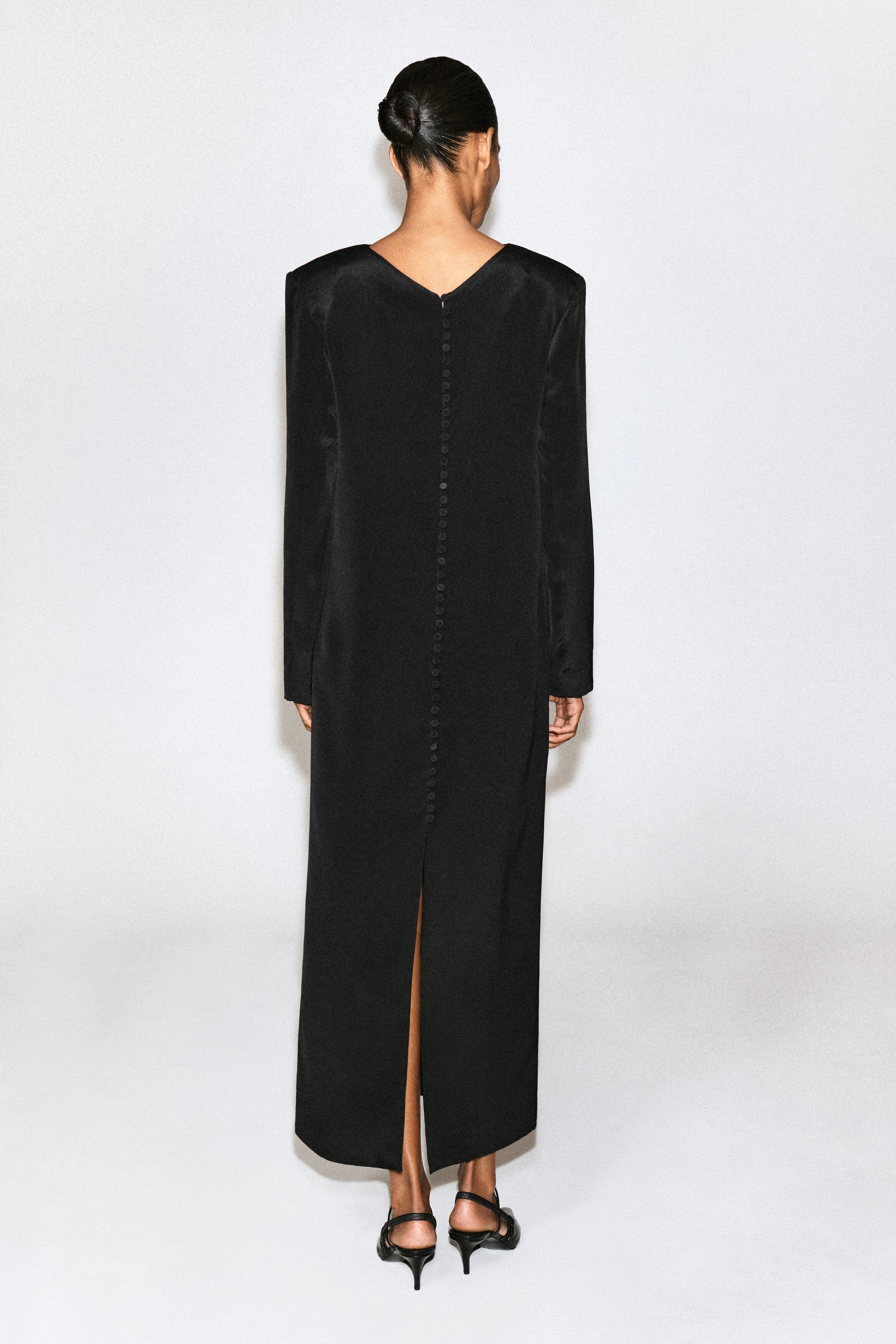 A black long-sleeved maxi dress featuring structured shoulders, a V-back neckline, a vertical row of fabric-covered buttons, and a centre-back slit is worn by the model. The dress is styled with black low-heeled sandals.