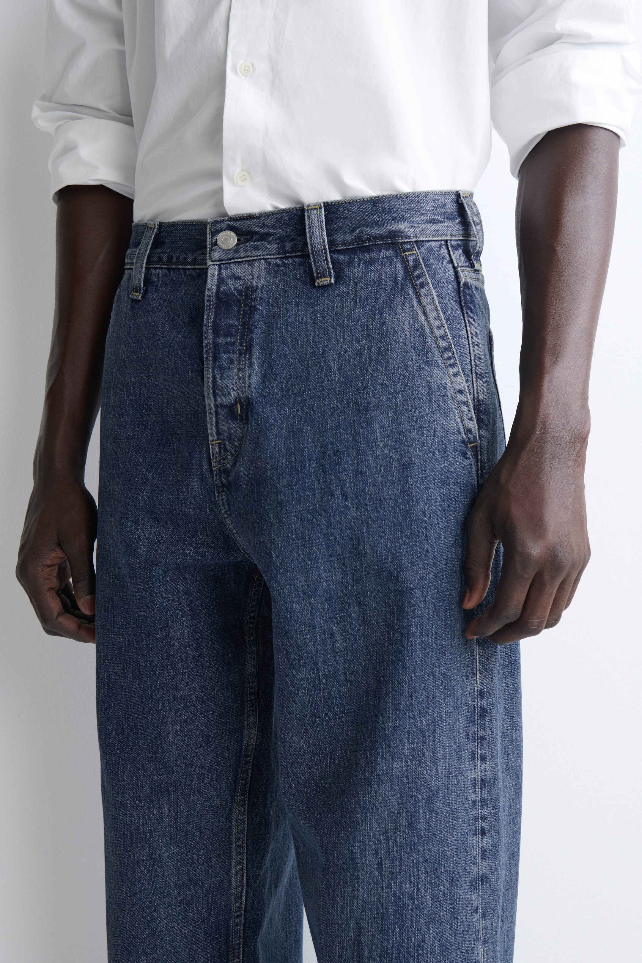 The relaxed-fit straight-leg denim trousers in a medium-blue wash are shown in close-up, featuring a high-rise waist with belt loops, a metal button fastening, and slanted side pockets. They are styled with a tucked-in white button-down shirt to clearly display the structured waistband and relaxed fit. The relaxed-fit straight-leg denim trousers in a medium-blue wash are shown in close-up, featuring a high-rise waist with belt loops, a metal button fastening, and slanted side pockets. They are styled with a tucked-in white button-down shirt to clearly display the structured waistband and relaxed fit.