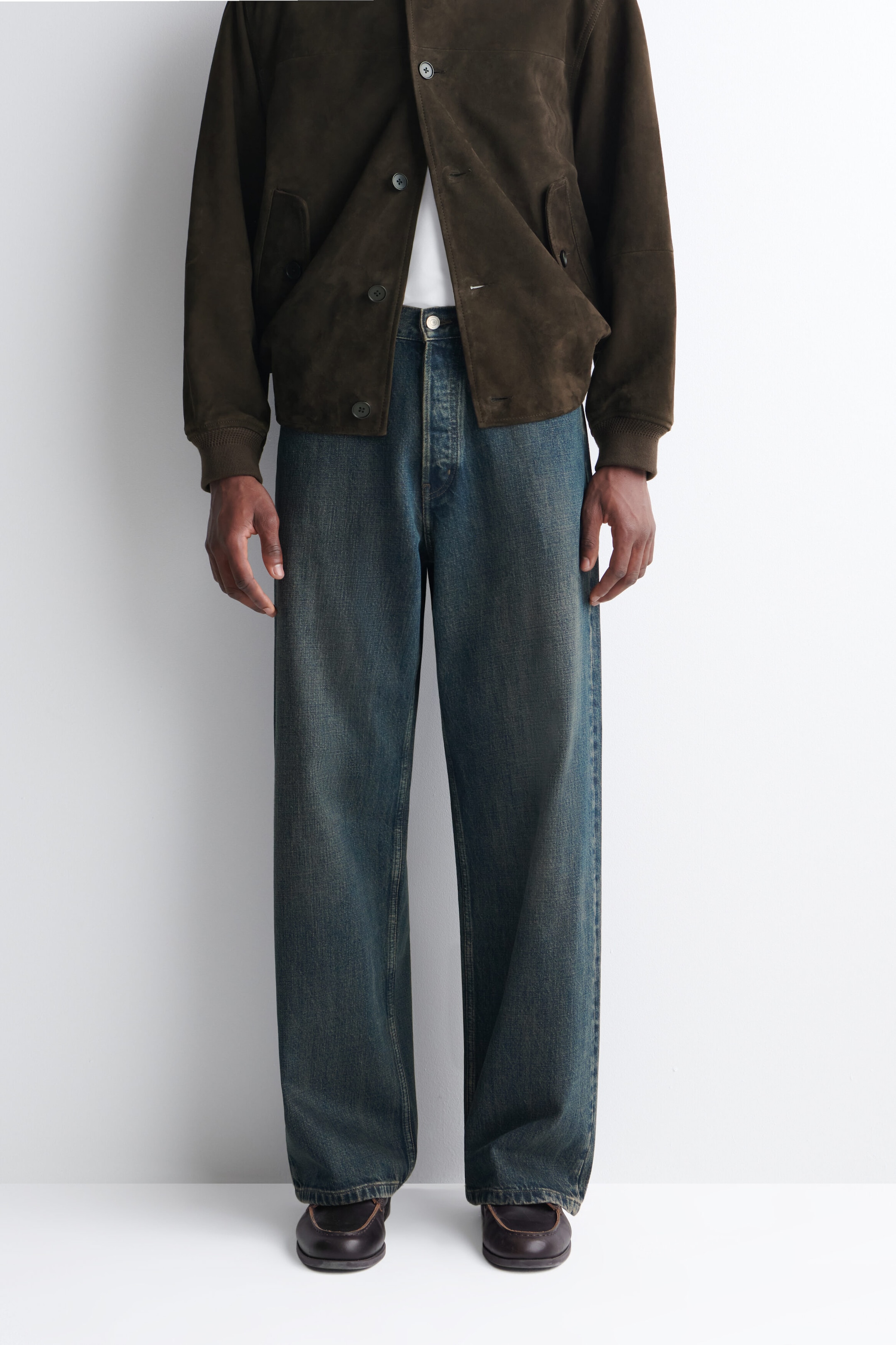 Wide-leg blue jeans with a faded wash are being worn by the model, styled with a partially buttoned brown suede jacket and dark brown leather shoes. Wide-leg blue jeans with a faded wash are being worn by the model, styled with a partially buttoned brown suede jacket and dark brown leather shoes.