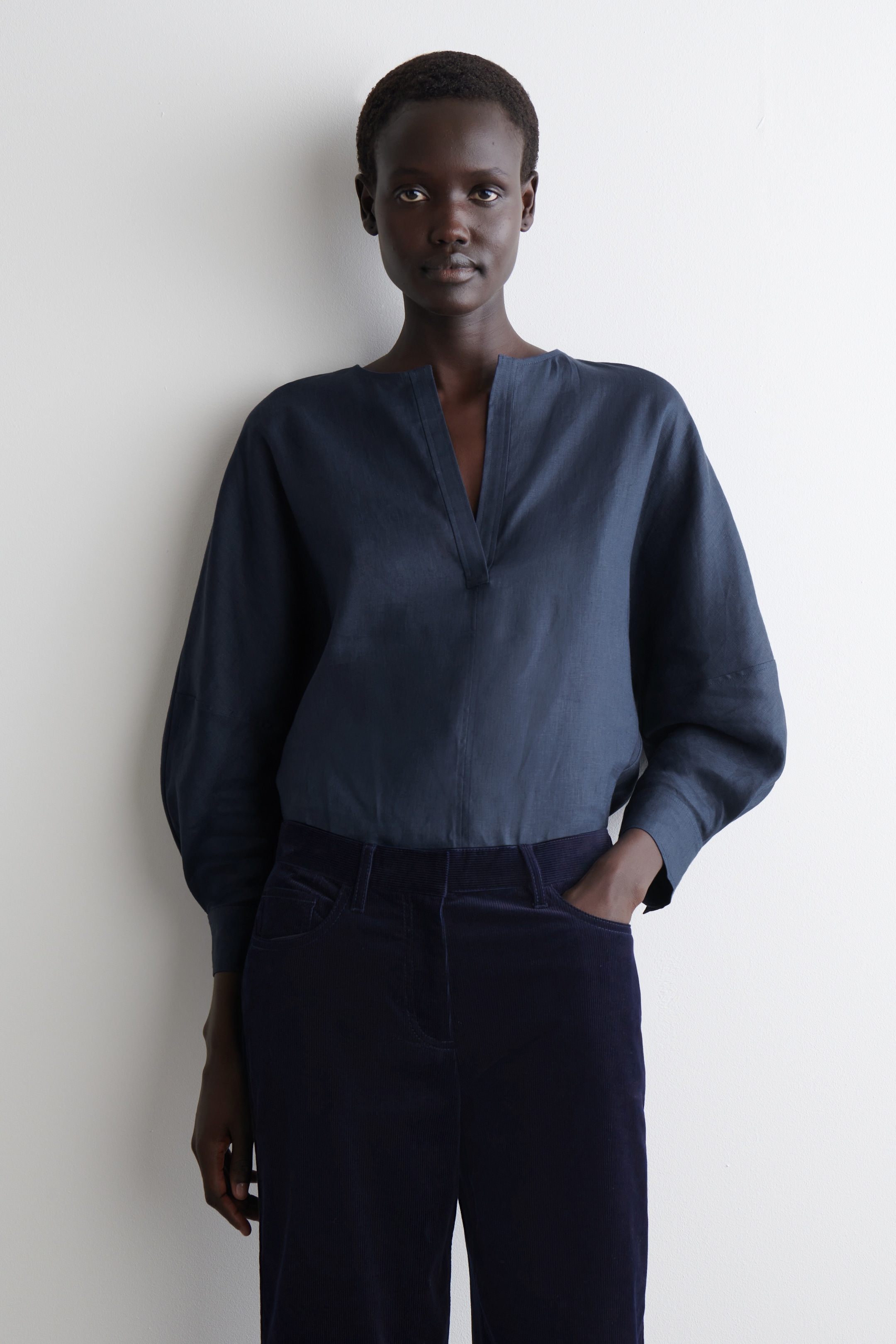 Visualizza immagine più grande: Un top blu scuro a vestibilità rilassata presenta uno scollo a V con fascia e maniche lunghe leggermente voluminose. È abbinato a pantaloni a gamba dritta in velluto a coste blu scuro, che presentano creste verticali visibili e tasche frontali.