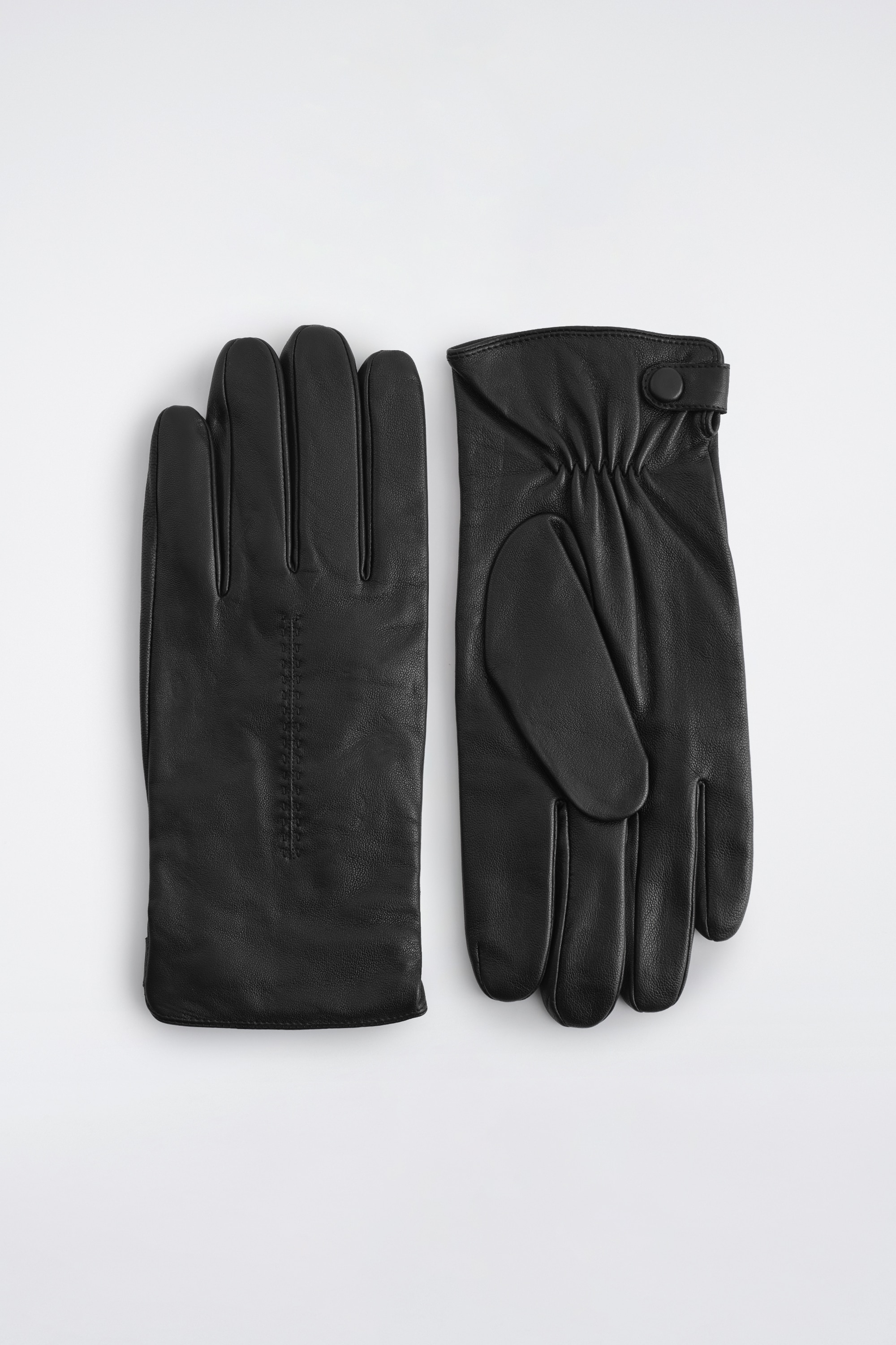Un gant en cuir noir est portÃ© Ã  la main tournÃ©e vers la gauche, mettant en valeur une texture lisse avec des dÃ©tails de piqÃ»res pintuck sur le dessus. Le gant est stylisÃ© glissÃ© sous la manchette d'une veste en cuir noir texturÃ©.