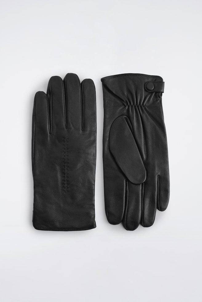 Leren handschoenen met kasjmiervoering liggen plat, waarbij één handschoen de achterkant van de hand toont en de andere de palmzijde. De handschoen links heeft een centrale verticale gestikte naad, terwijl de handschoen rechts een elastische pols met een bandje met drukknoopsluiting heeft. - 1