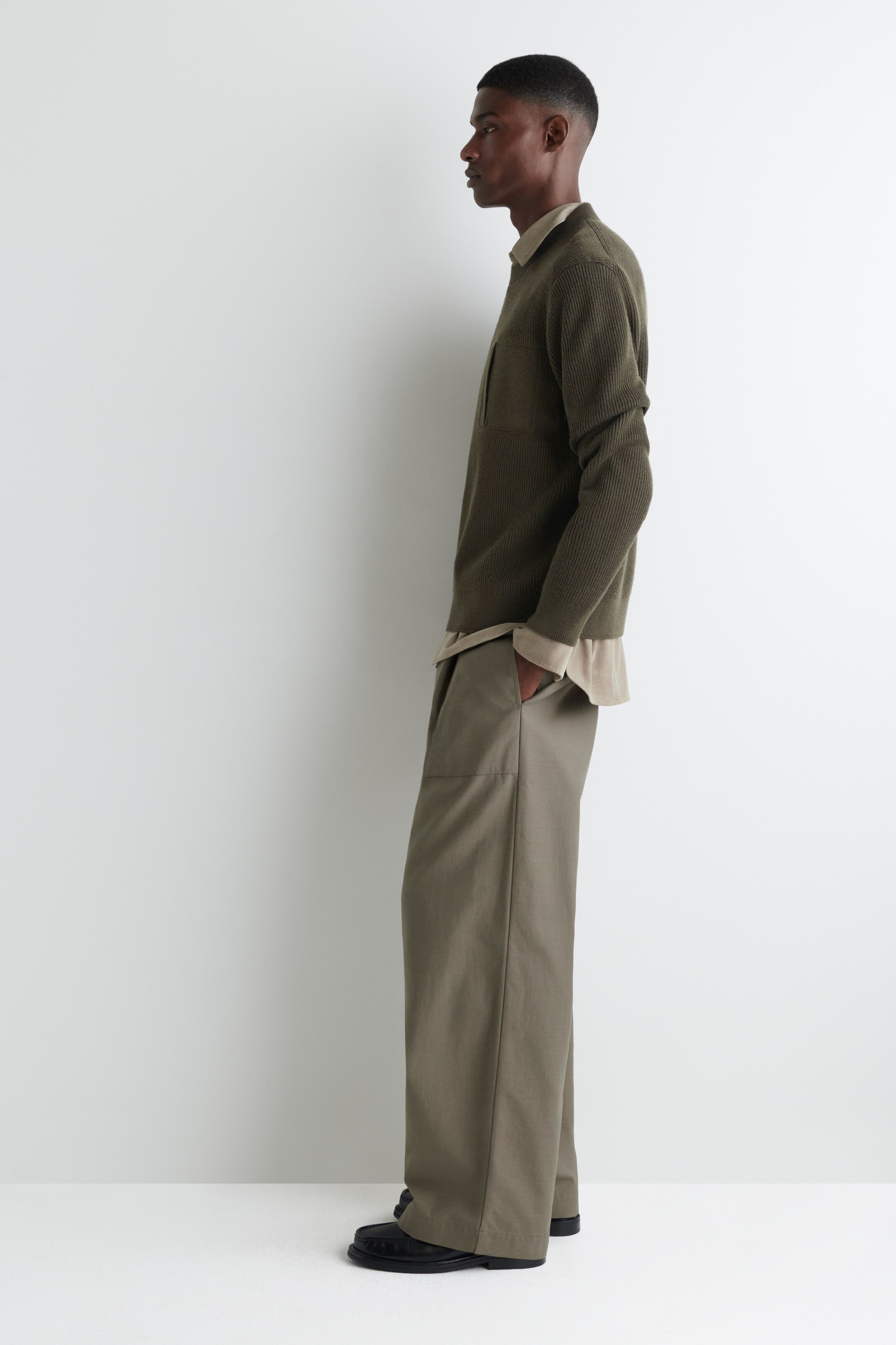 Agrandir l'image: Un haut polo à manches longues côtelé de couleur olive foncée, avec une poche poitrine et un col doux, est superposé à une chemise beige clair à col, visible au niveau du cou et des poignets. Un pantalon kaki ample à jambes larges complète le look, avec des détails de coutures visibles sur le devant des jambes, accompagné de chaussures noires, le tout présenté en profil latéral.