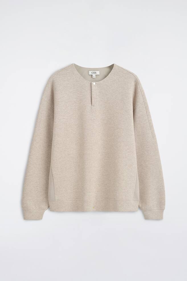 Eine Frontansicht des lÃ¤ssigen Scuba-Henley-Tops zeigt einen hellbeigen Melange-Stoff mit Rundhalsausschnitt und einer kurzen Knopfleiste mit einem einzelnen Druckknopfverschluss. Das Design prÃ¤sentiert Ã¼berschnittene Schultern, lange Ãrmel mit RippbÃ¼ndchen und einen passenden gerippten Saum. - 1